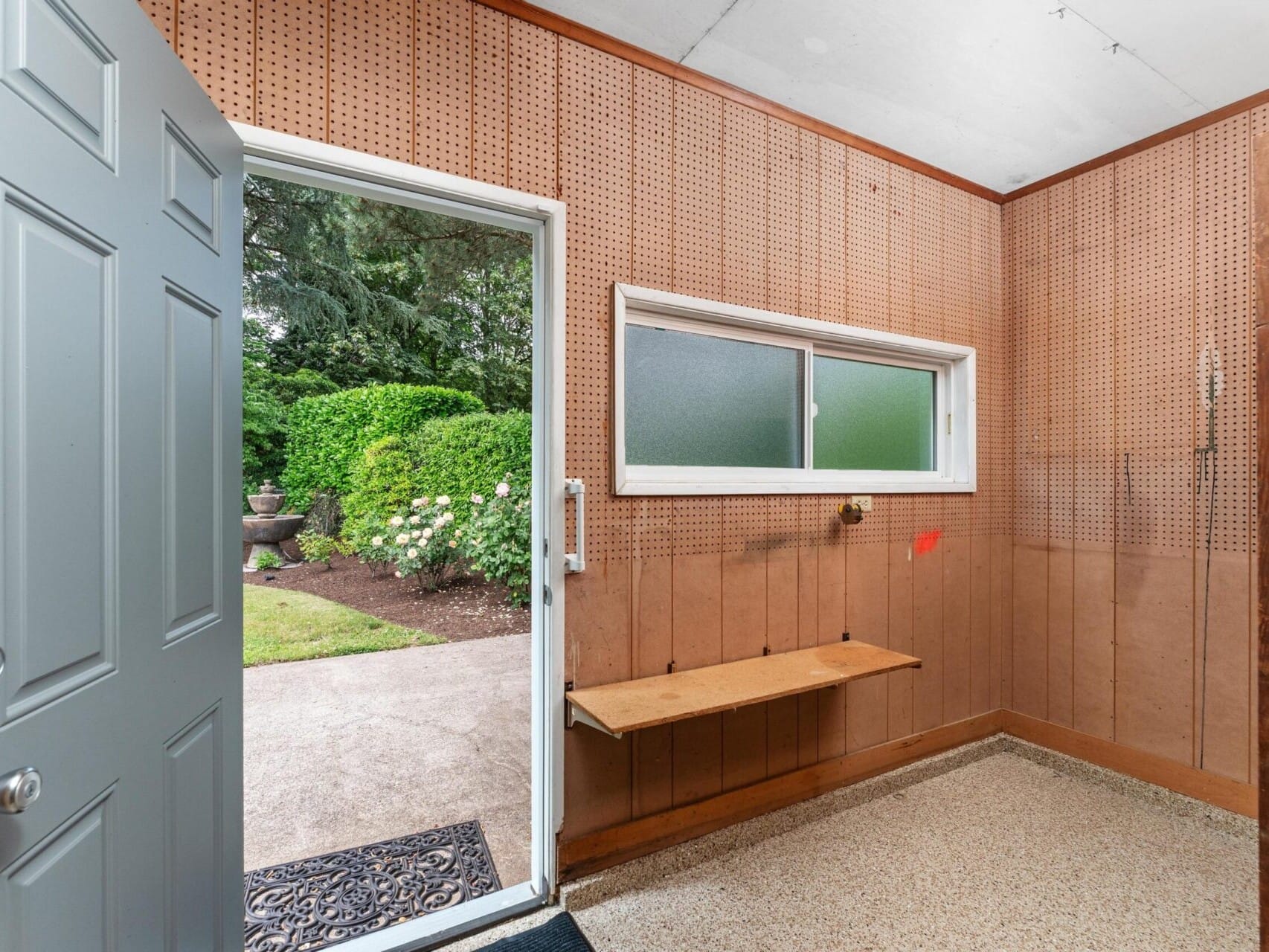 A small room with pegboard walls, a wooden shelf, and an open gray door leading outside. The floor is speckled, and a rectangular window has frosted glass. Outside, there are lush green bushes and a stone vase with flowers.