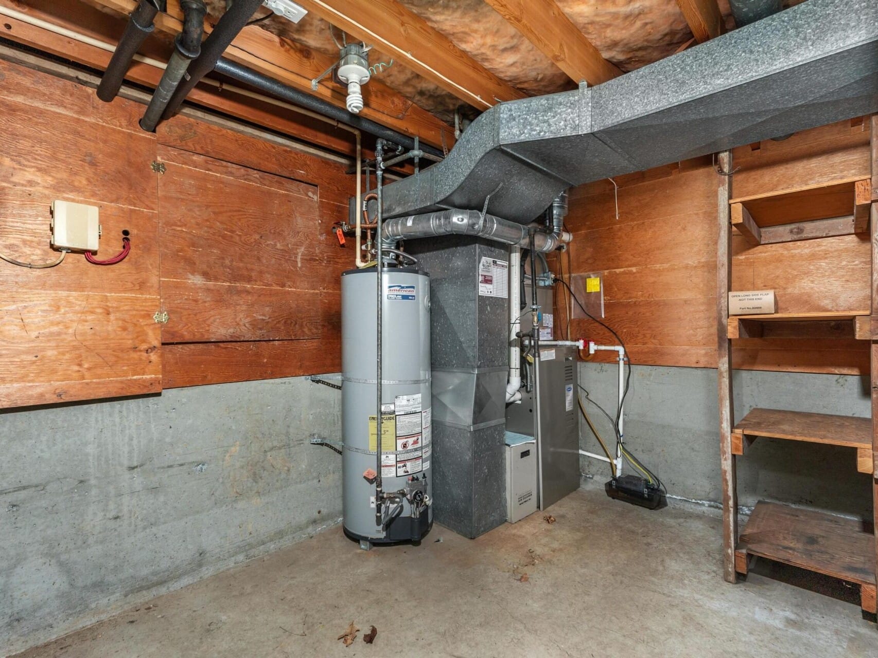 Image of a basement with an exposed ceiling and wooden walls. It contains a water heater, furnace, and ductwork. Wooden shelves are on the right side, and wires and pipes run across the walls and ceiling. The floor is concrete.