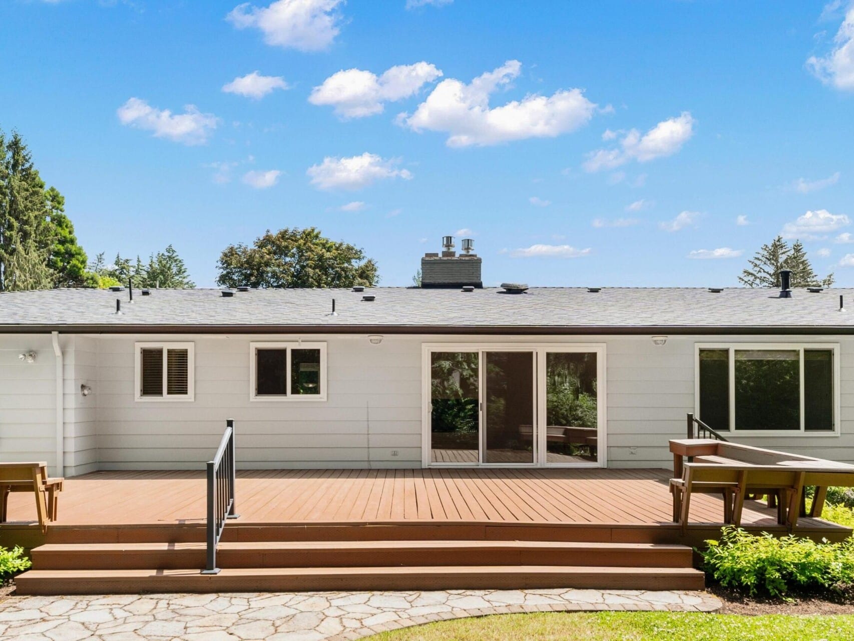 A modern single-story house with white siding, large glass doors, and a wooden deck. The yard is landscaped with greenery and a stone path. The sky is clear with a few clouds.