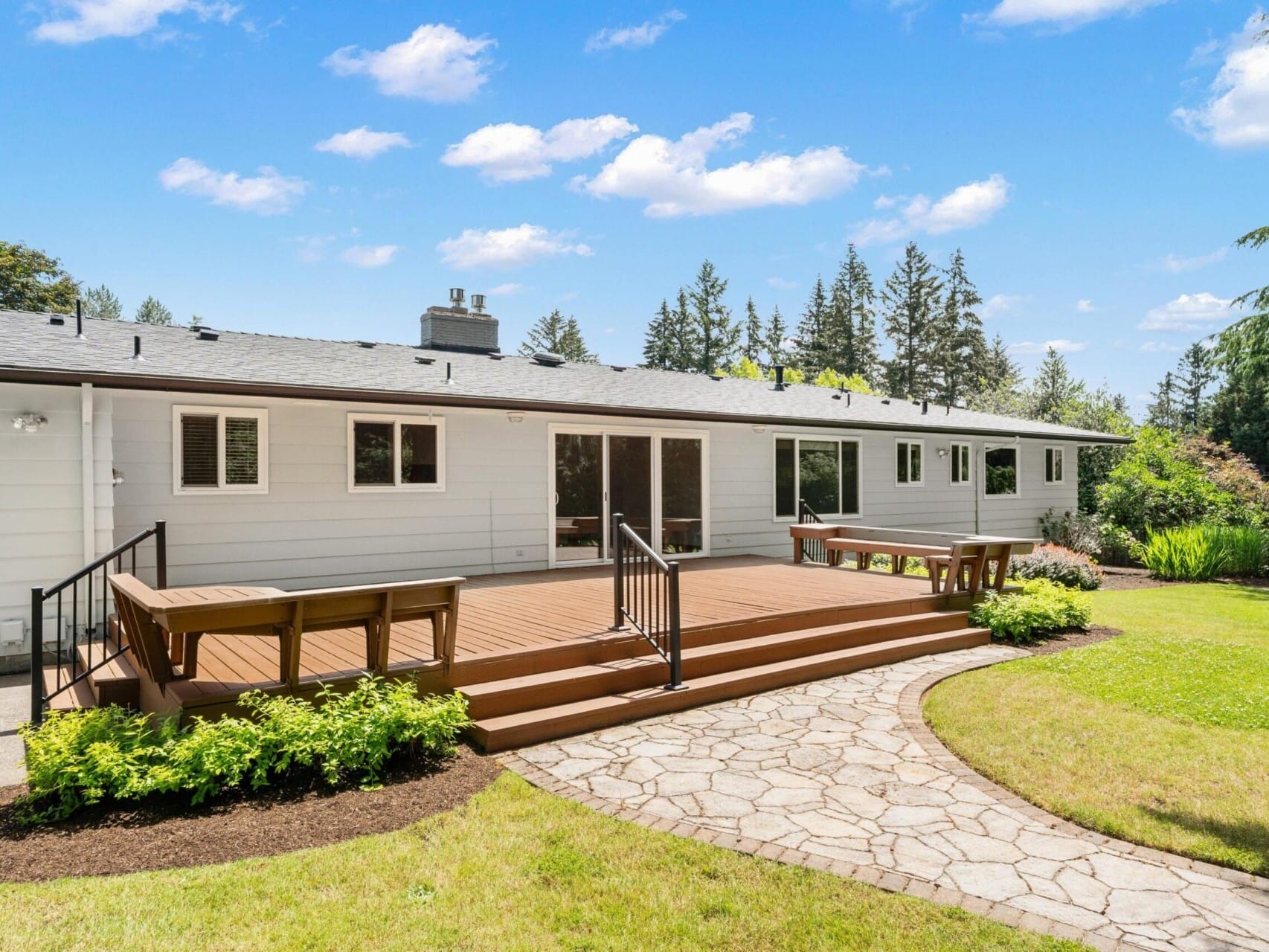 A spacious backyard featuring a wooden deck with benches, surrounded by a stone pathway and lush green grass. The house has large windows and is set against a backdrop of tall trees under a clear blue sky with scattered clouds.