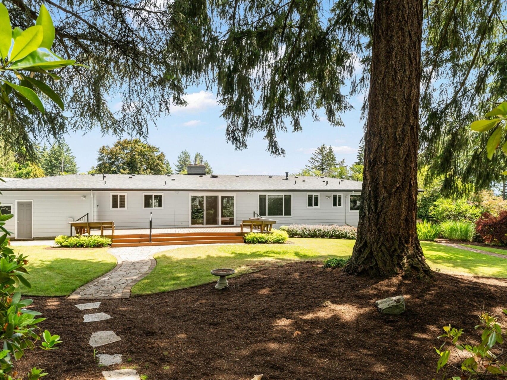 A white single-story house with large windows stands in a lush backyard. Theres a wooden deck and stone path leading to the house. Tall trees and various plants surround the yard, adding greenery under a clear blue sky.