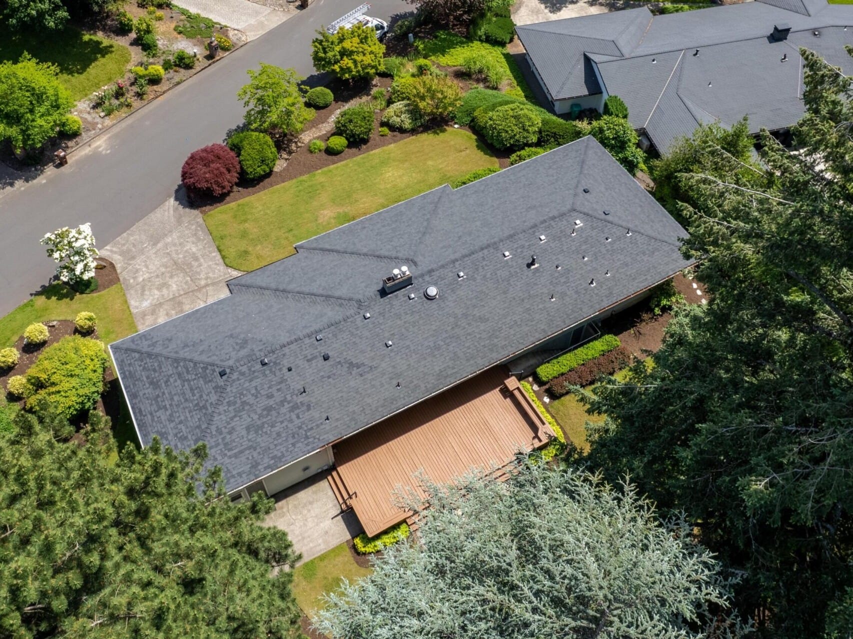 Aerial view of a suburban house with a gray roof and a wooden deck. The property is surrounded by lush greenery, trees, and shrubs, with a driveway leading to the road. Other houses are visible nearby.