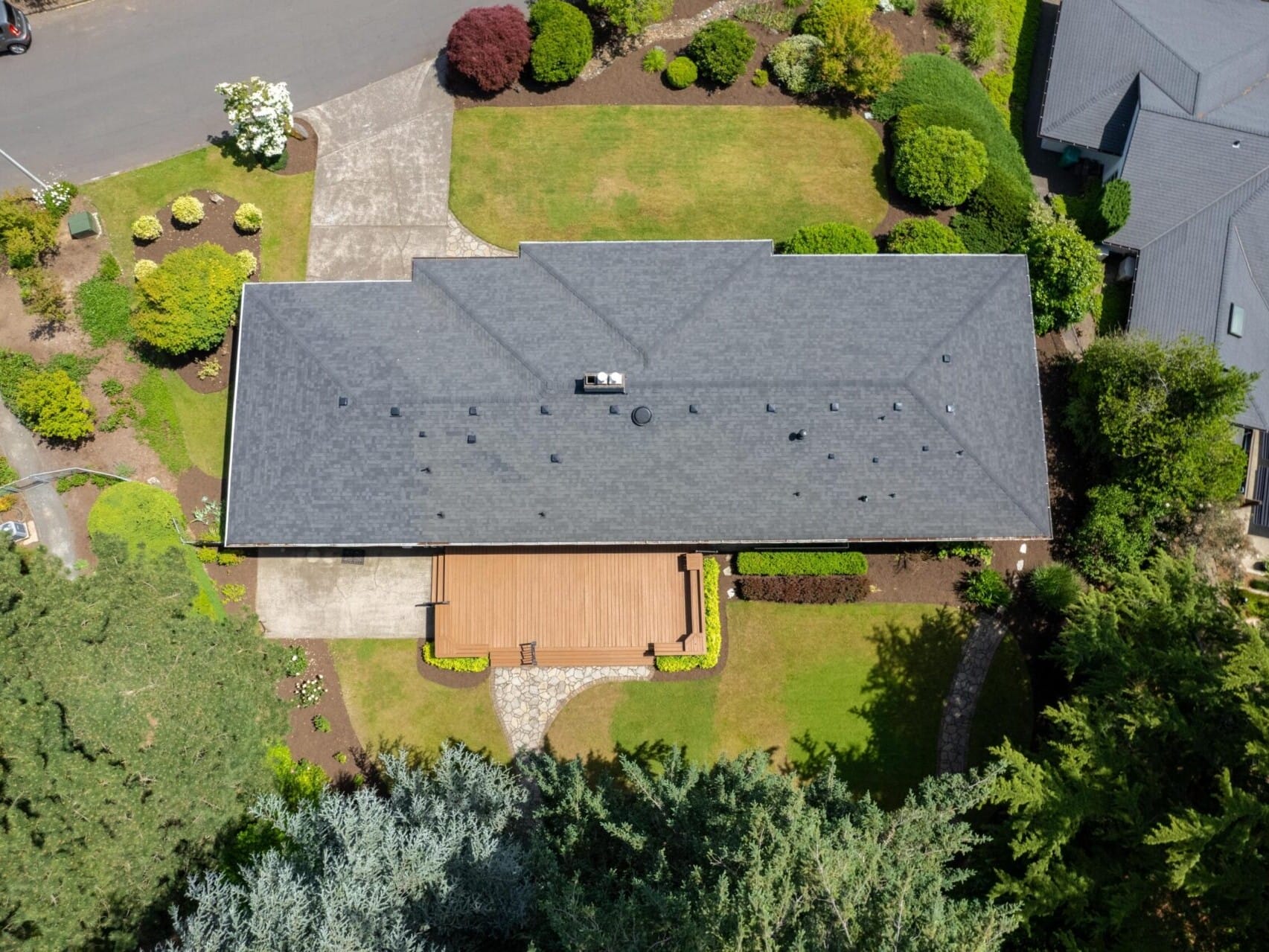 Aerial view of a house with a gray roof, surrounded by well-maintained gardens. A large wooden deck extends from the house, and there are trees and shrubs in the yard. A road runs beside the property.