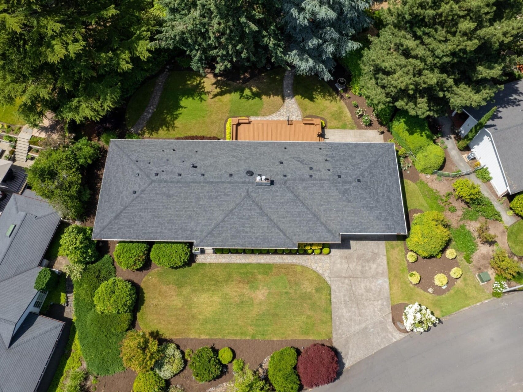 Aerial view of a house with a gray roof, surrounded by a well-maintained garden and pathway. The backyard features a wooden deck. The property is flanked by neighboring houses and lush green trees.