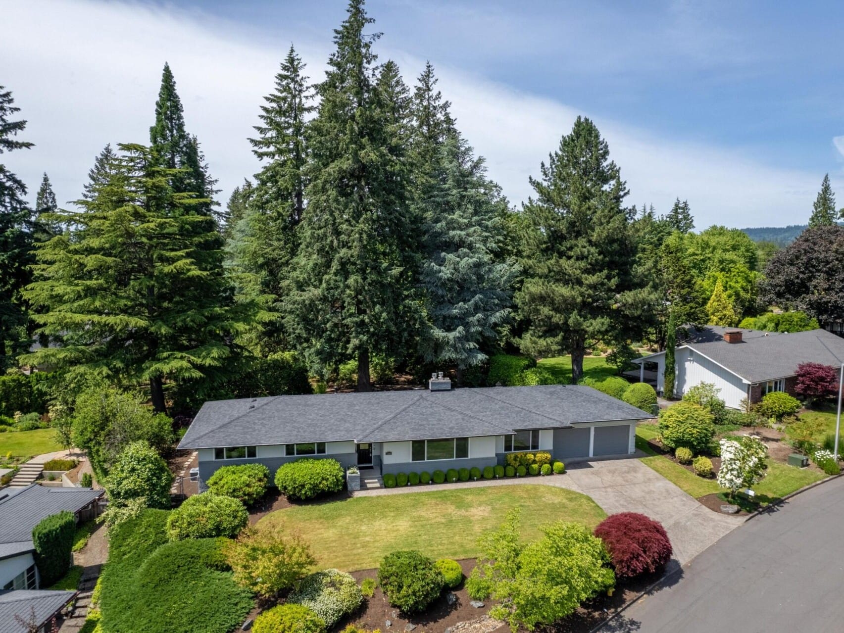 Aerial view of a suburban house surrounded by lush greenery and trees. The single-story home has a gray roof and is set on a well-maintained lawn with bushes. A driveway leads to the house, and neighboring homes are visible on a curved street.