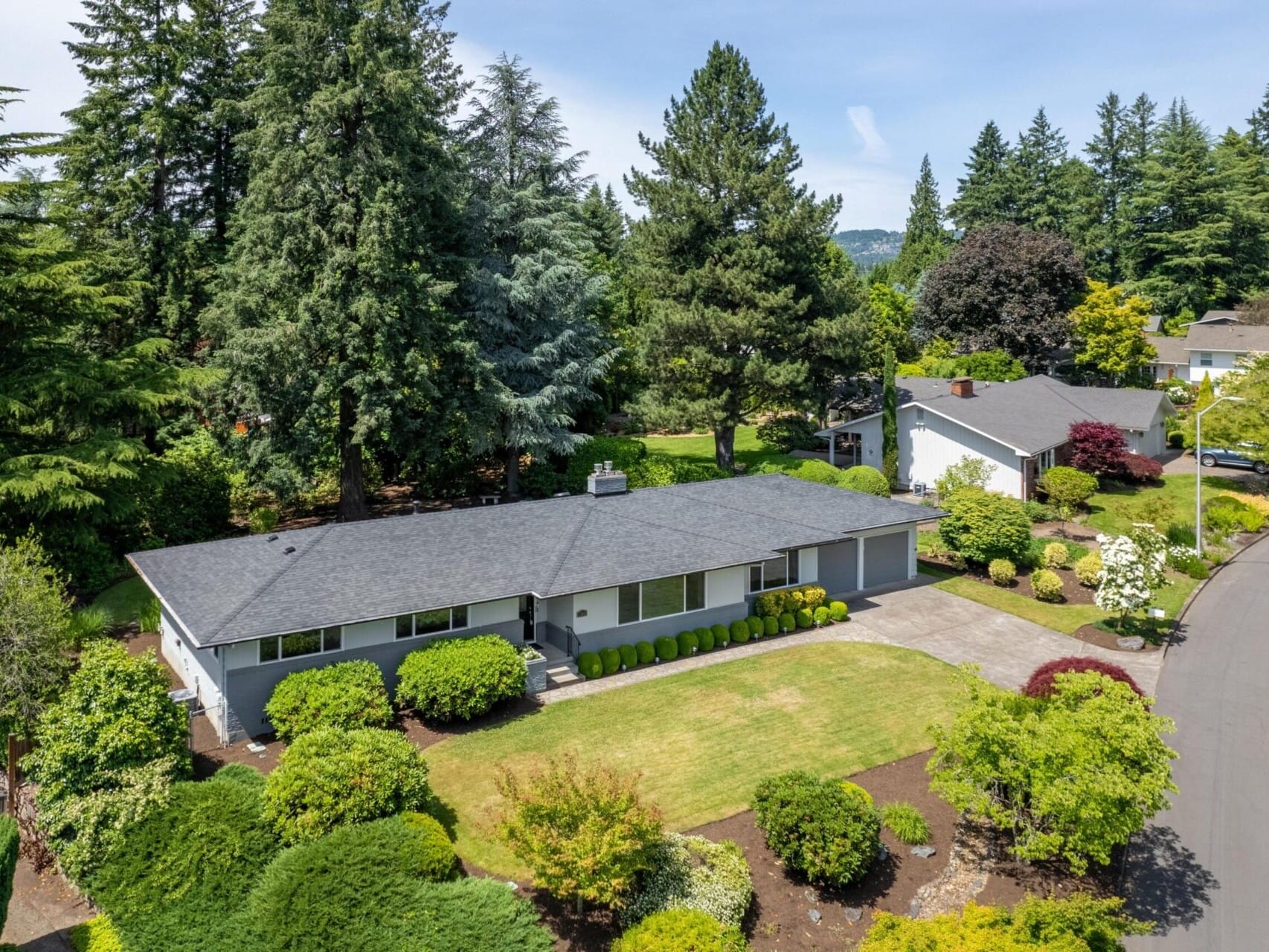 Aerial view of a suburban house surrounded by lush greenery and tall trees. The house has a dark roof and a well-maintained lawn with neatly trimmed bushes. A driveway leads to a garage, and the street curves gently beside the property.