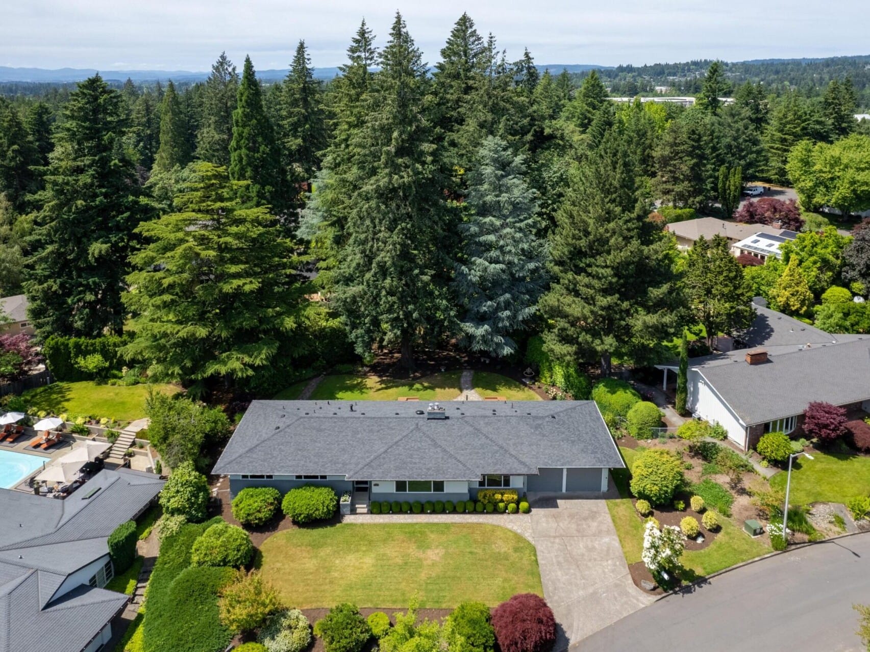 Aerial view of a residential neighborhood with a single-story house surrounded by lush greenery and tall trees. The property features a manicured lawn and garden. Nearby, other homes and a backyard with a pool are visible.