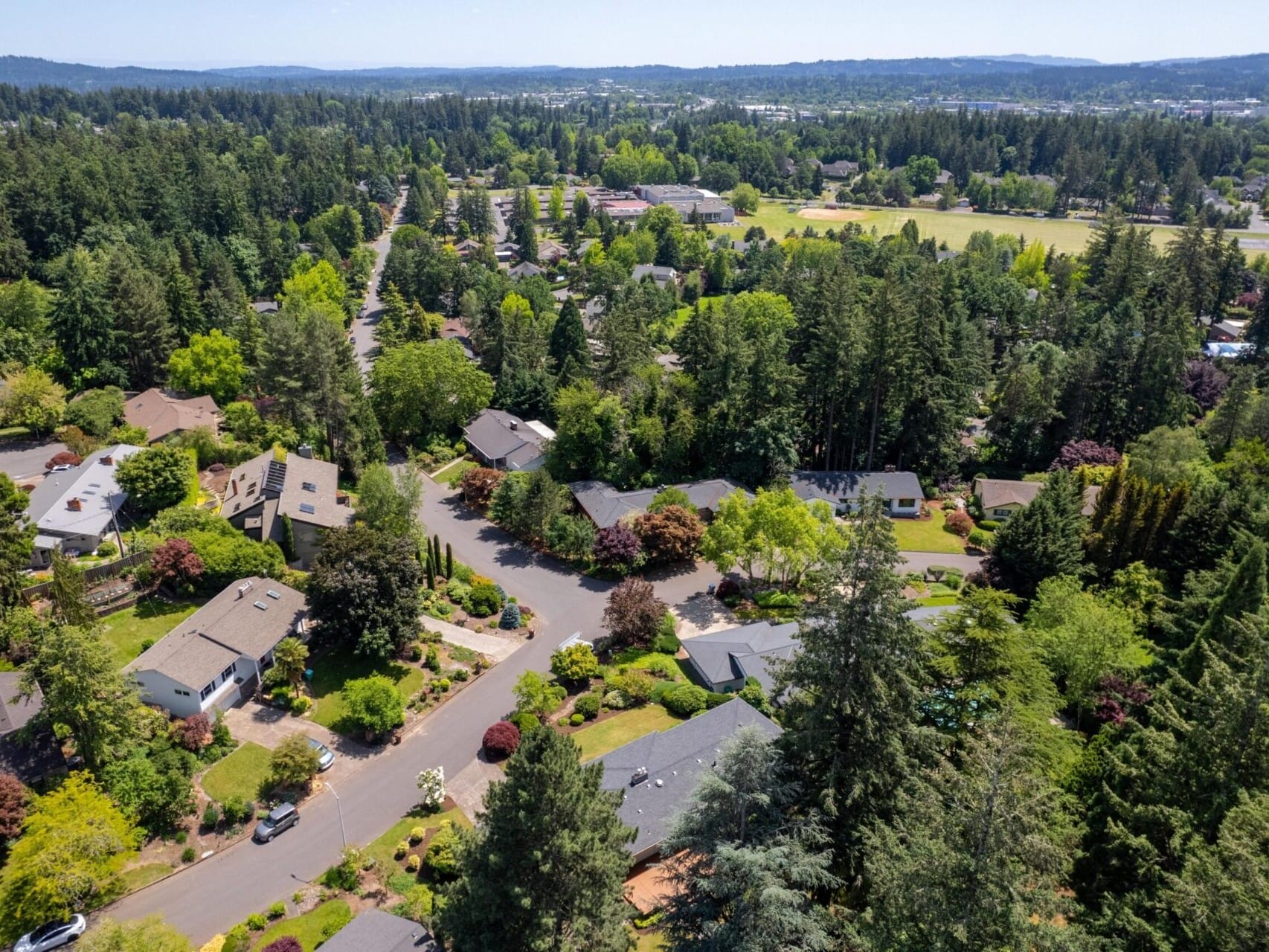 Aerial view of a suburban neighborhood surrounded by lush greenery and trees. Houses with yards and streets are visible, set against a backdrop of rolling hills and distant horizons under a clear blue sky.