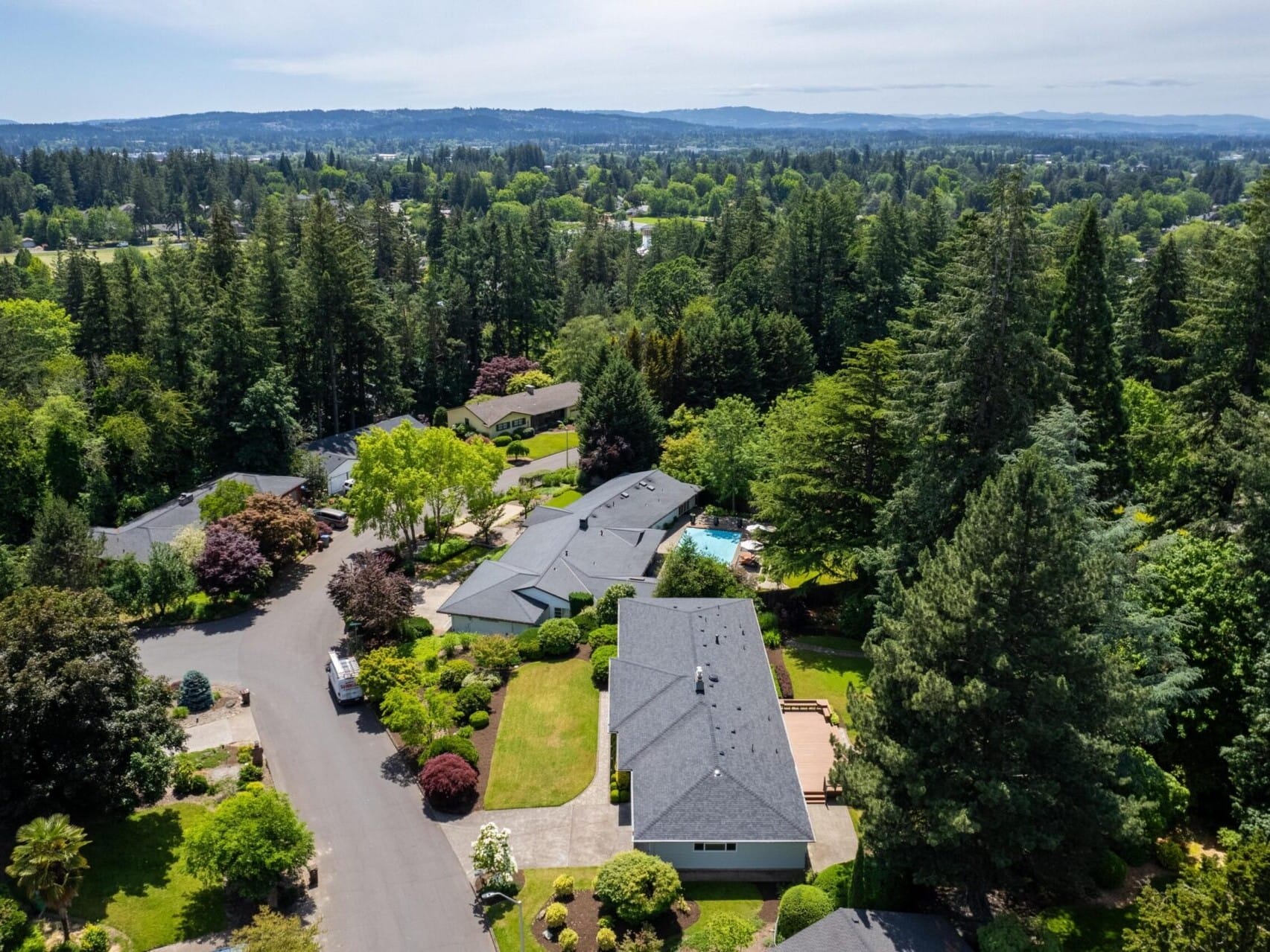 Aerial view of a residential neighborhood surrounded by dense green forests. The scene shows several houses with grey roofs, well-maintained lawns, and a circular driveway. In the background, a vast expanse of trees and distant hills is visible under a cloudy sky.