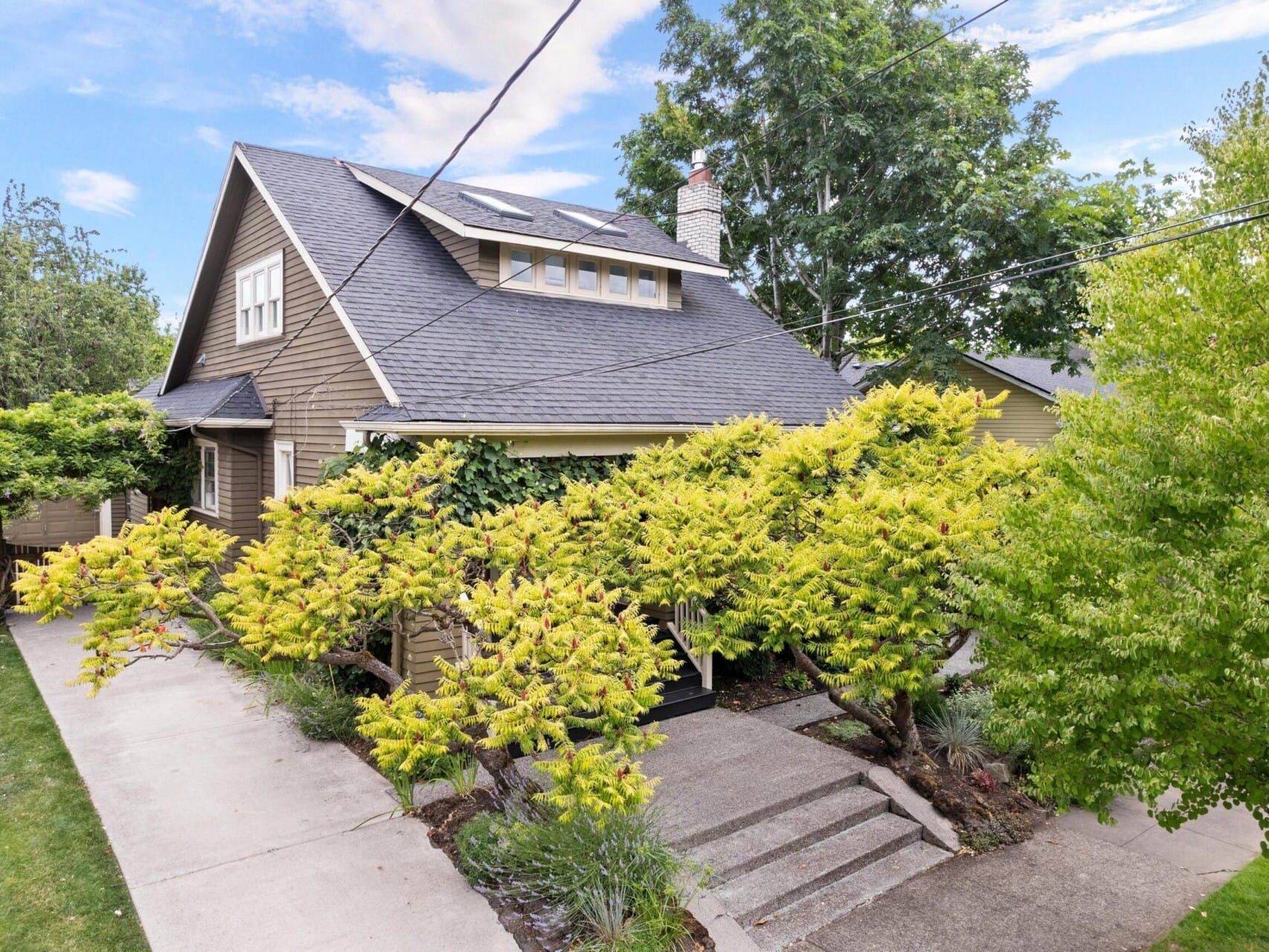 A charming house with a steep roof and a chimney, nestled among lush green trees and shrubs. A concrete pathway leads to the entrance, under Portland's partly cloudy sky. Discover this gem with a top Portland Realtor today.