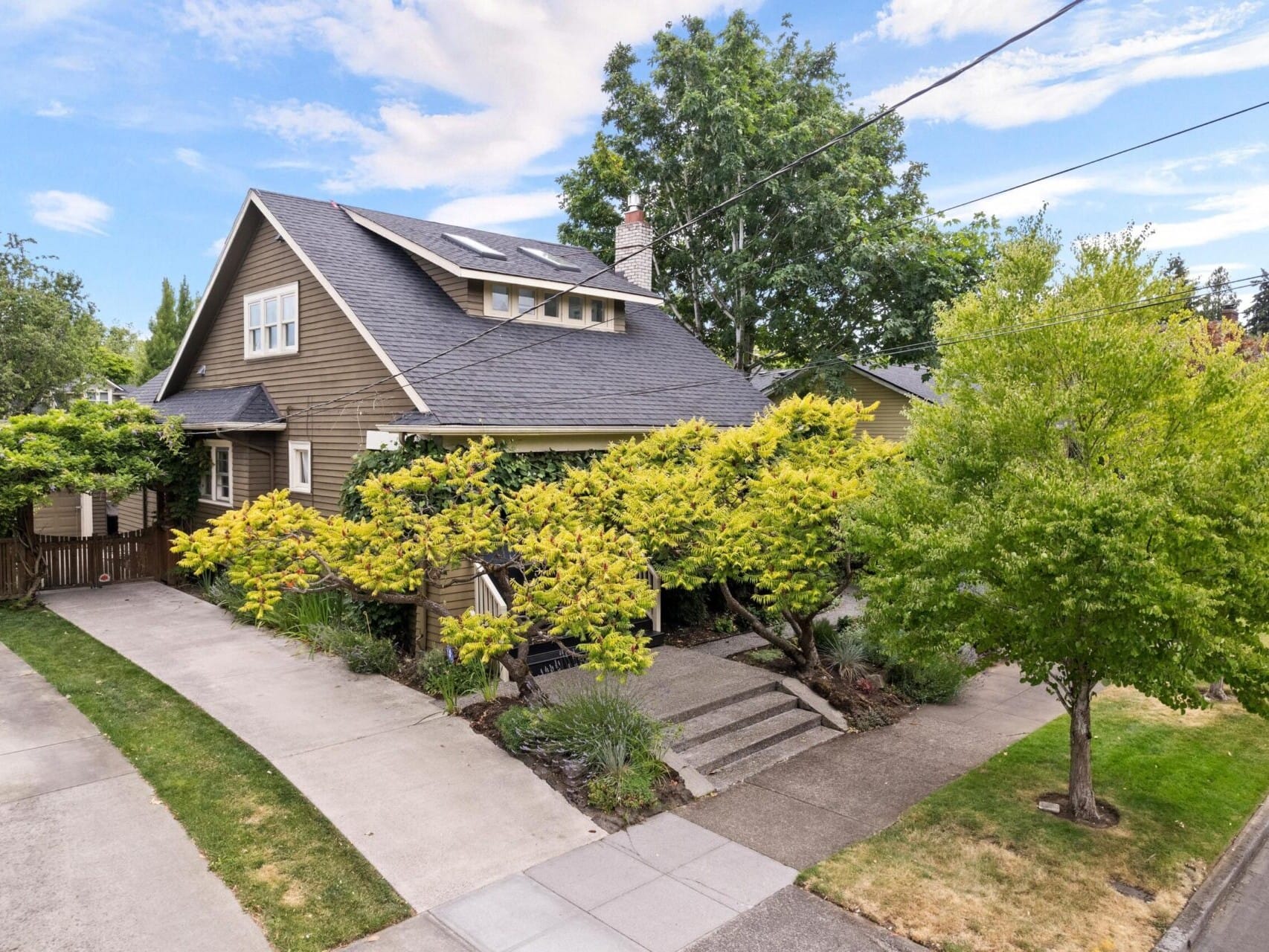 A charming, two-story house with a gabled roof, nestled among lush green trees and shrubs. This Portland Oregon real estate gem features a wooden exterior and a paved driveway leading to a small path with steps. The sky is partly cloudy, enhancing the serene atmosphere.