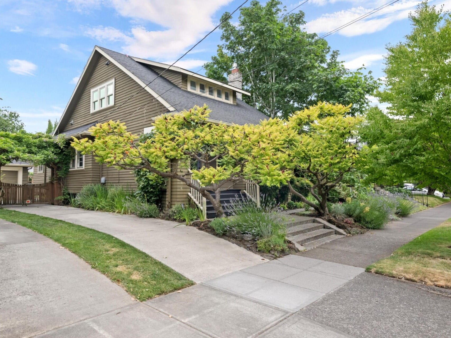 A charming house with a steep, black roof and tan siding, surrounded by lush greenery and trees, reflects the serene beauty of Portland Oregon real estate. The wide driveway and pathway invite you to an inviting entrance under partly cloudy, blue skies.