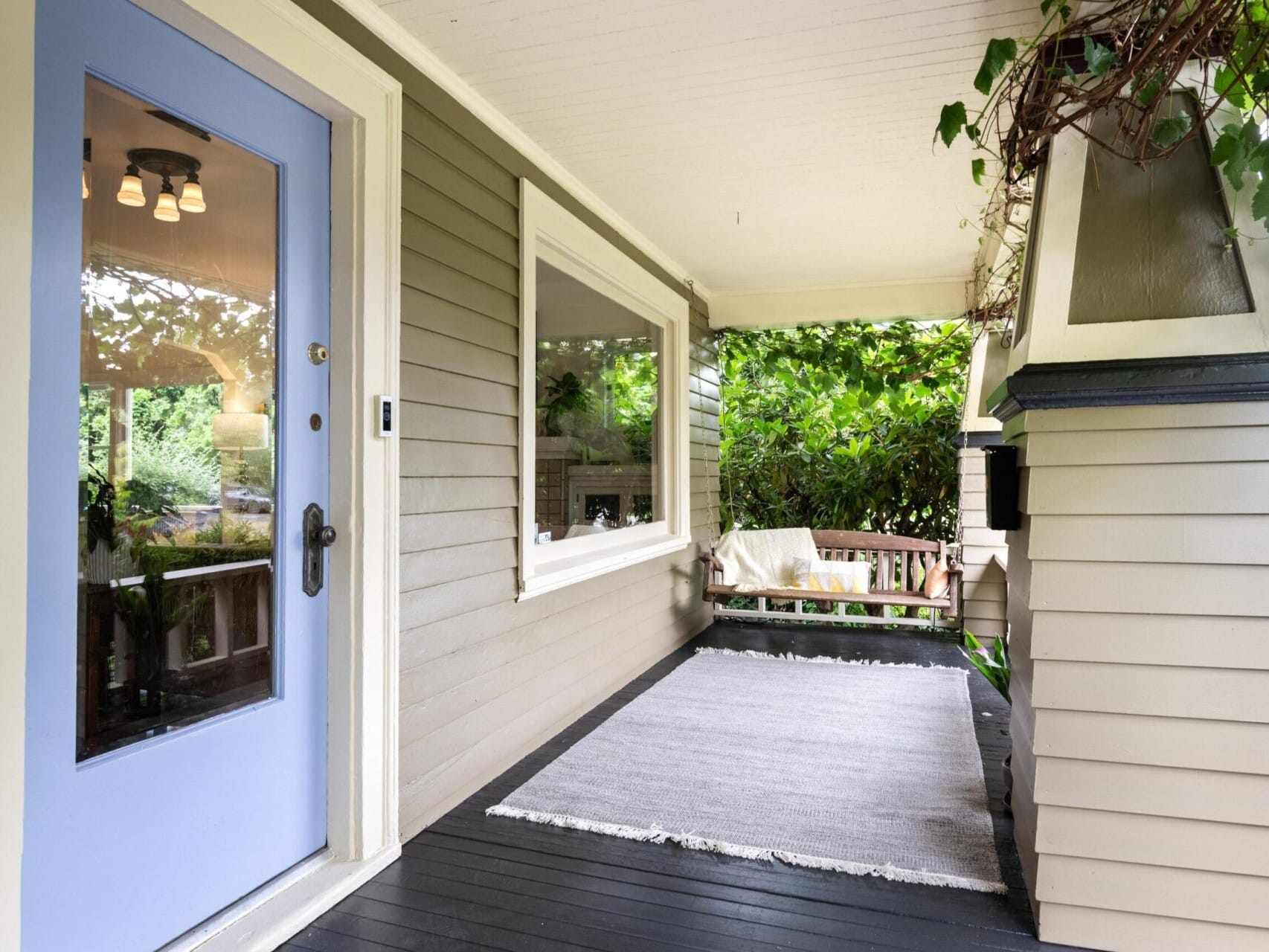 A cozy porch featuring a light blue door, a large window, and a hanging porch light is the epitome of Portland Oregon real estate charm. A wooden bench with a blanket sits next to lush greenery, while an outdoor rug graces the dark wooden floorboards.