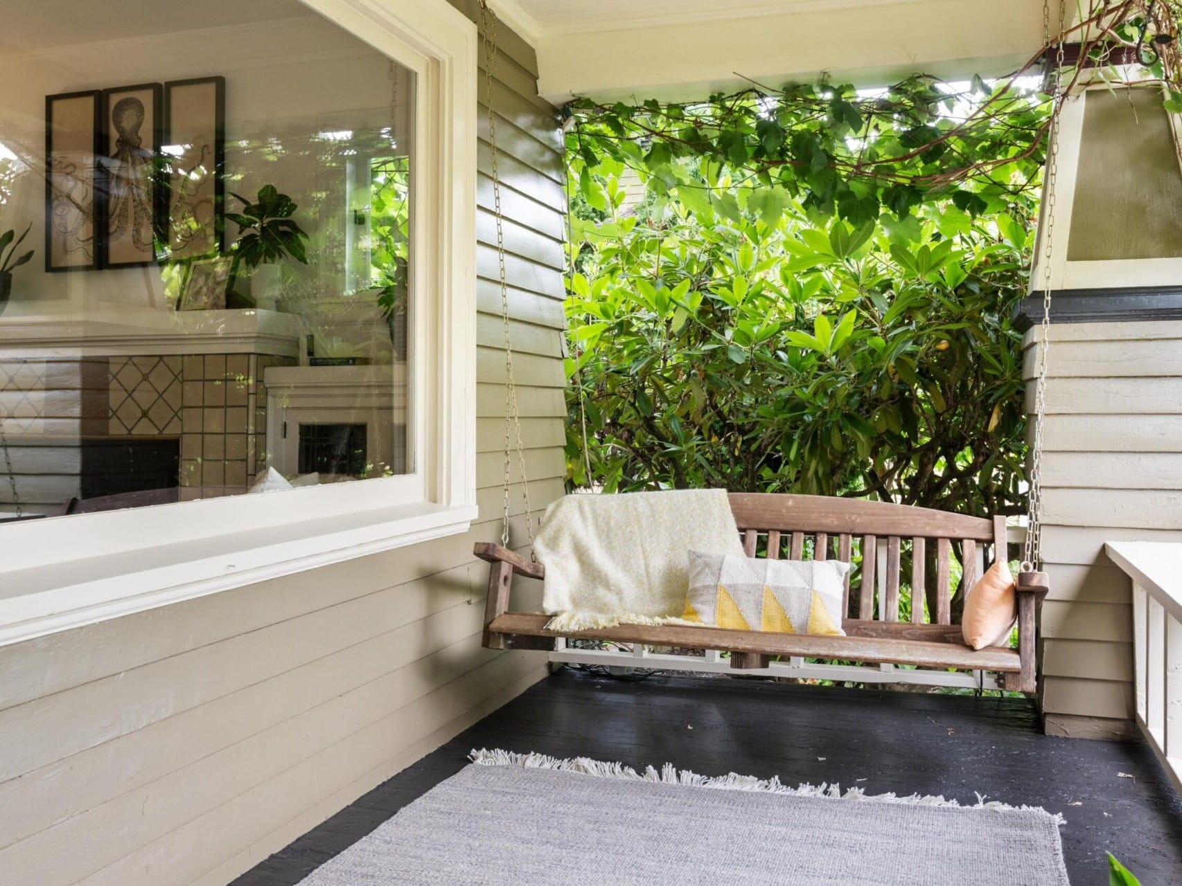 A cozy porch in Portland, Oregon, features a wooden swing with cushions and a blanket, surrounded by lush greenery. Attached to a house with a window showcasing interior decor, this charming space is part of the allure you'll find in Portland real estate. A gray rug covers part of the dark floor.