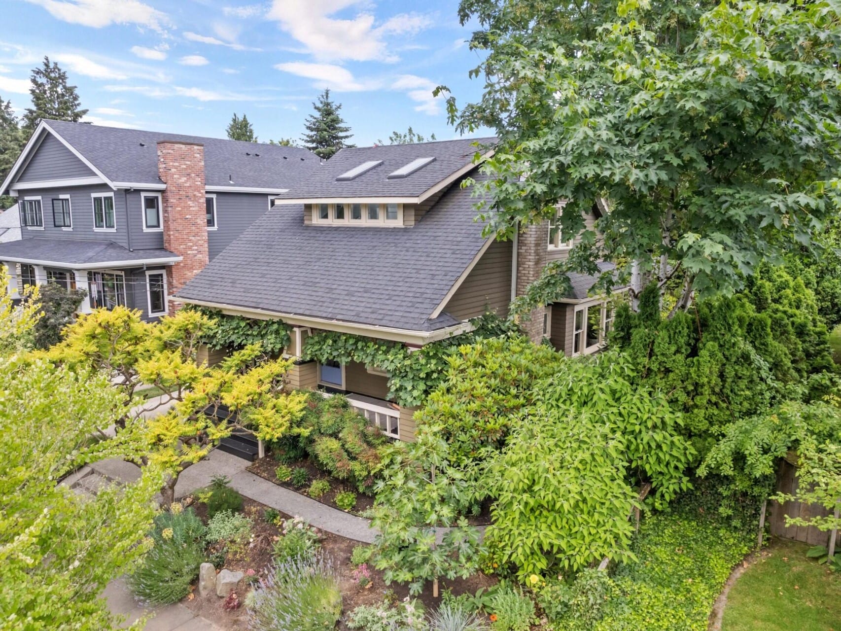 Aerial view of a charming two-story house in Portland, Oregon, with a gray roof nestled among lush green trees and shrubs. The home boasts skylights and large windows. A paved walkway winds through the front garden, offering a glimpse into desirable Portland real estate.