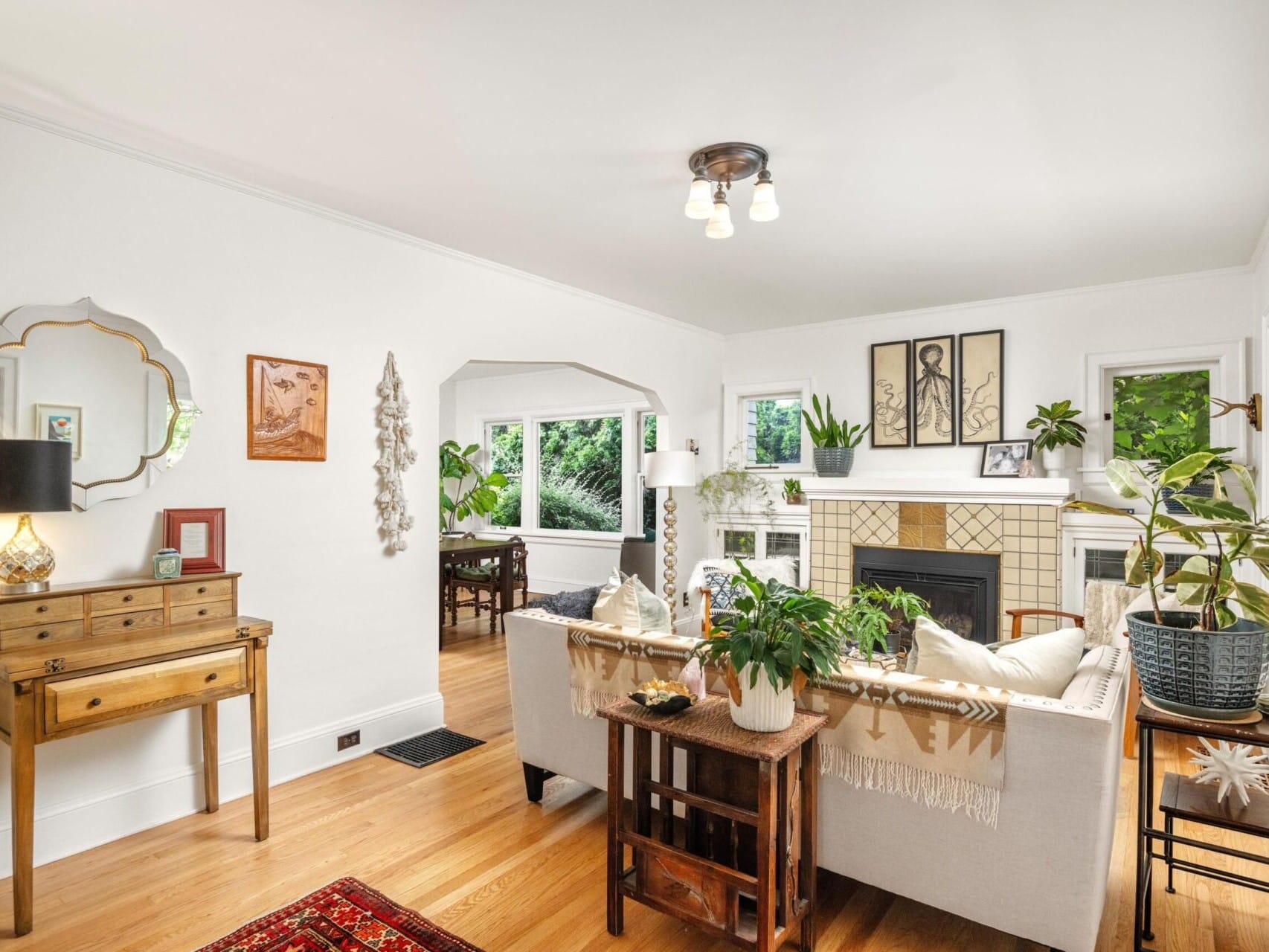 A cozy living room with a beige sofa, green plants, and a fireplace adorned with art. Wooden floors complement the modern-rustic mix of furniture, including a console table and round mirror. Large windows provide natural light and greenery views—perfect for any Portland Realtor showcasing charm.