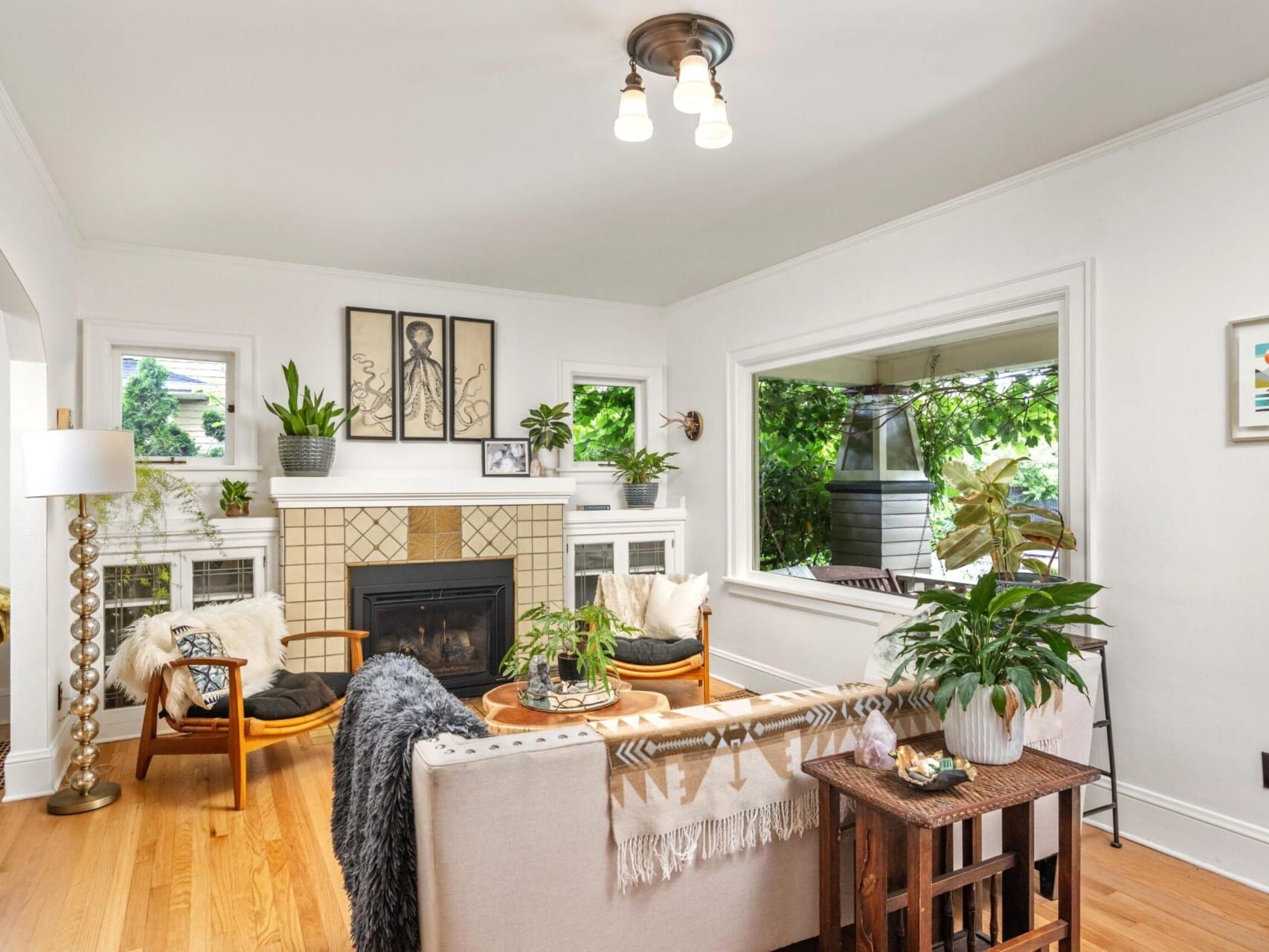 A cozy living room in a Portland Oregon real estate gem features a beige sofa, wooden floor, and fireplace. Adorned with plants, artwork, and a standing lamp, large windows offer a view of greenery outside, bathing the space in natural light.