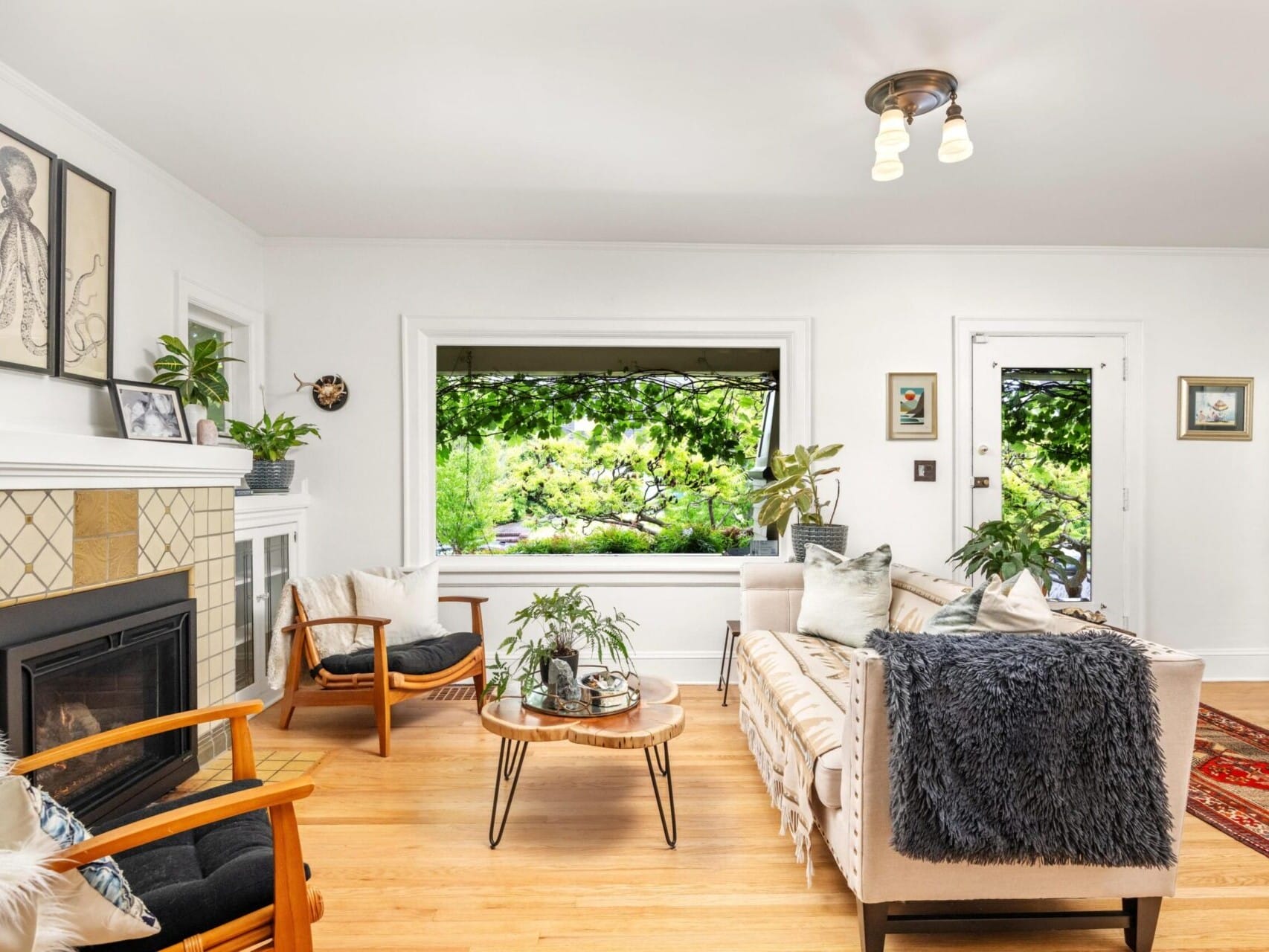 A cozy living room with a beige sofa, armchairs, and a wooden coffee table. The fireplace is adorned with plants and art, reflecting the charm that draws people to Portland Oregon real estate. Large windows offer views of lush greenery, complementing the light wood flooring and inviting rug below.