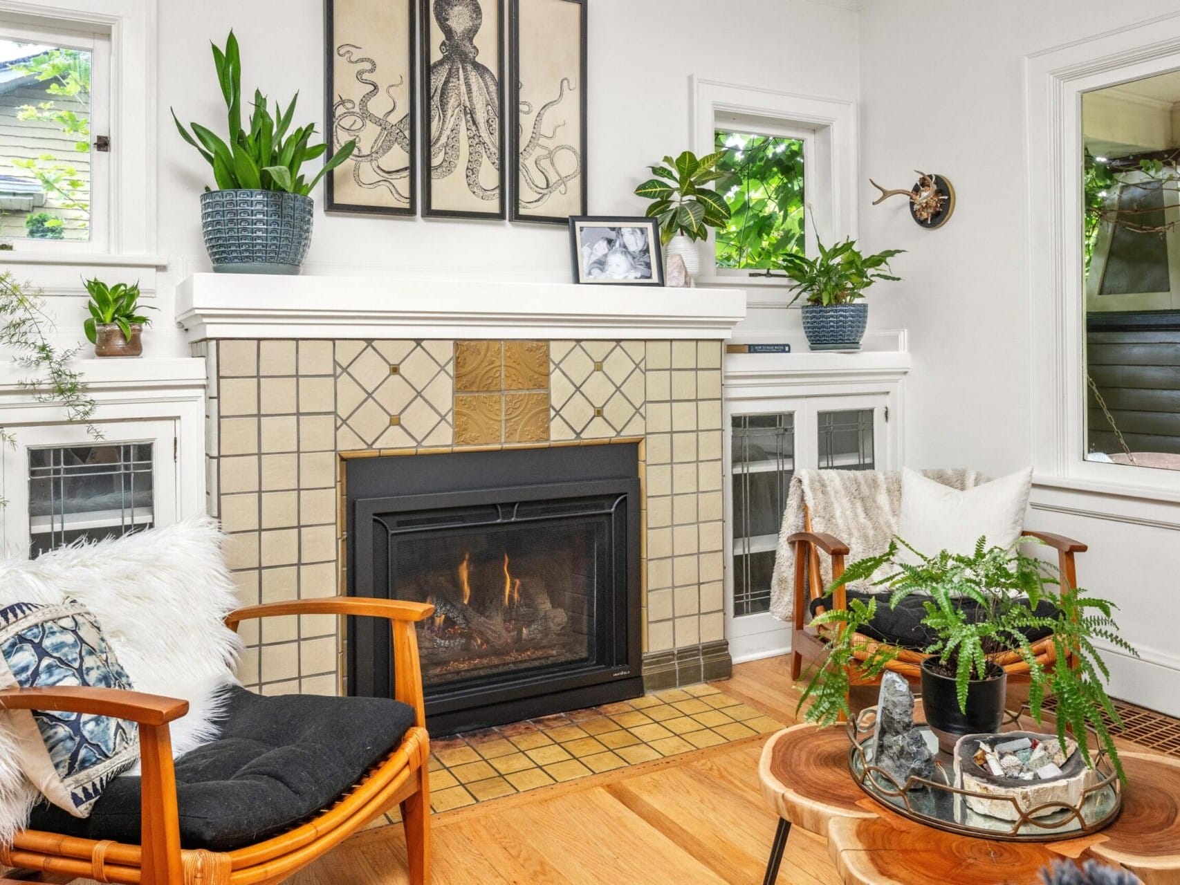 A cozy living room in desirable Portland Oregon real estate features a fireplace surrounded by tan tiles. Two wooden chairs with black cushions and fur accents face the fire. Plants adorn the mantel and side tables, while a large window displays outdoor greenery, complemented by octopus artwork above.