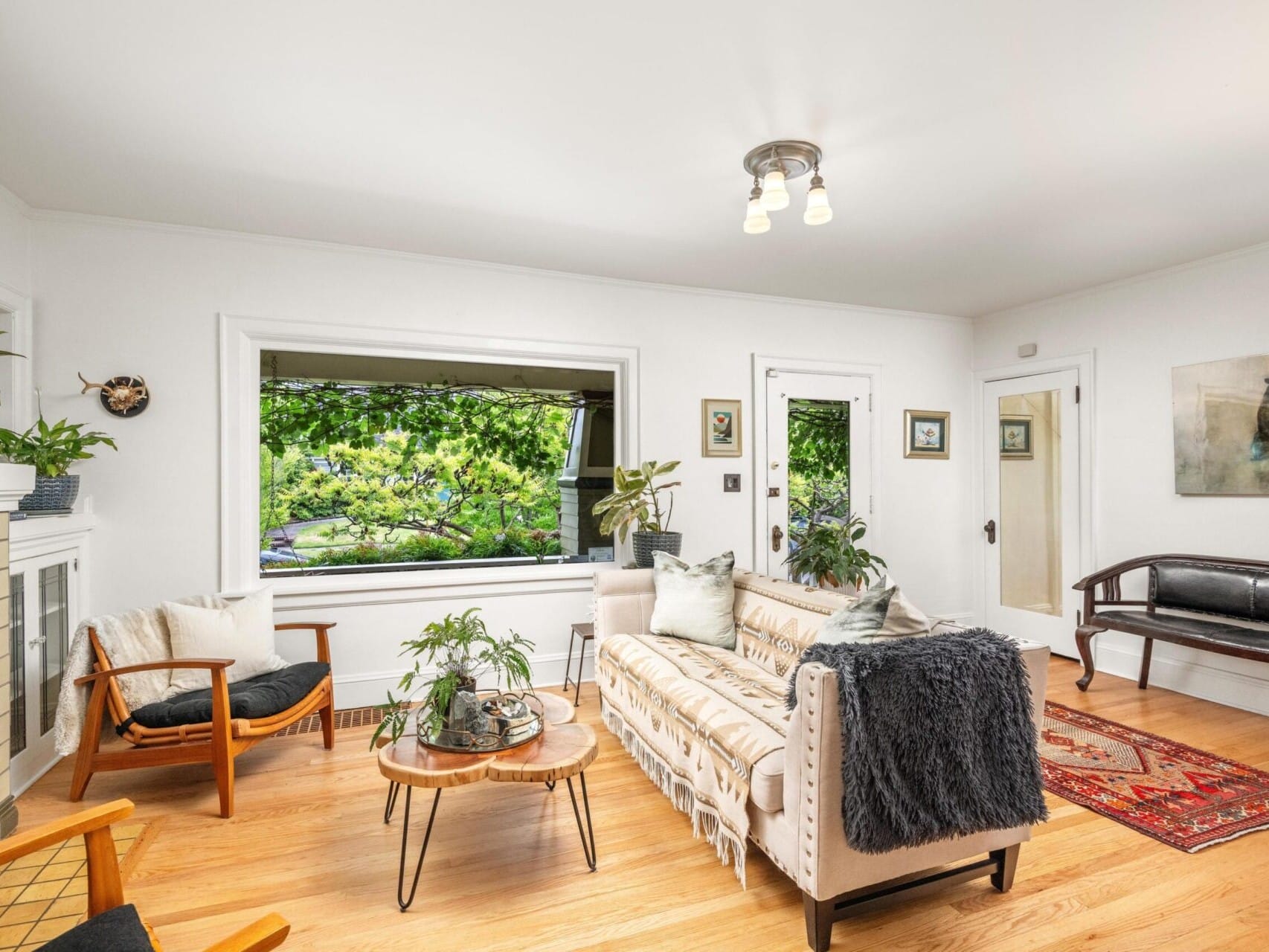 A cozy living room in a Portland Oregon real estate gem, showcasing a large window with lush greenery. It features a beige couch, chairs, fireplace, plants, and art. A wooden floor and colorful rug complete this inviting space. Perfect for those seeking the expertise of a top real estate agent.