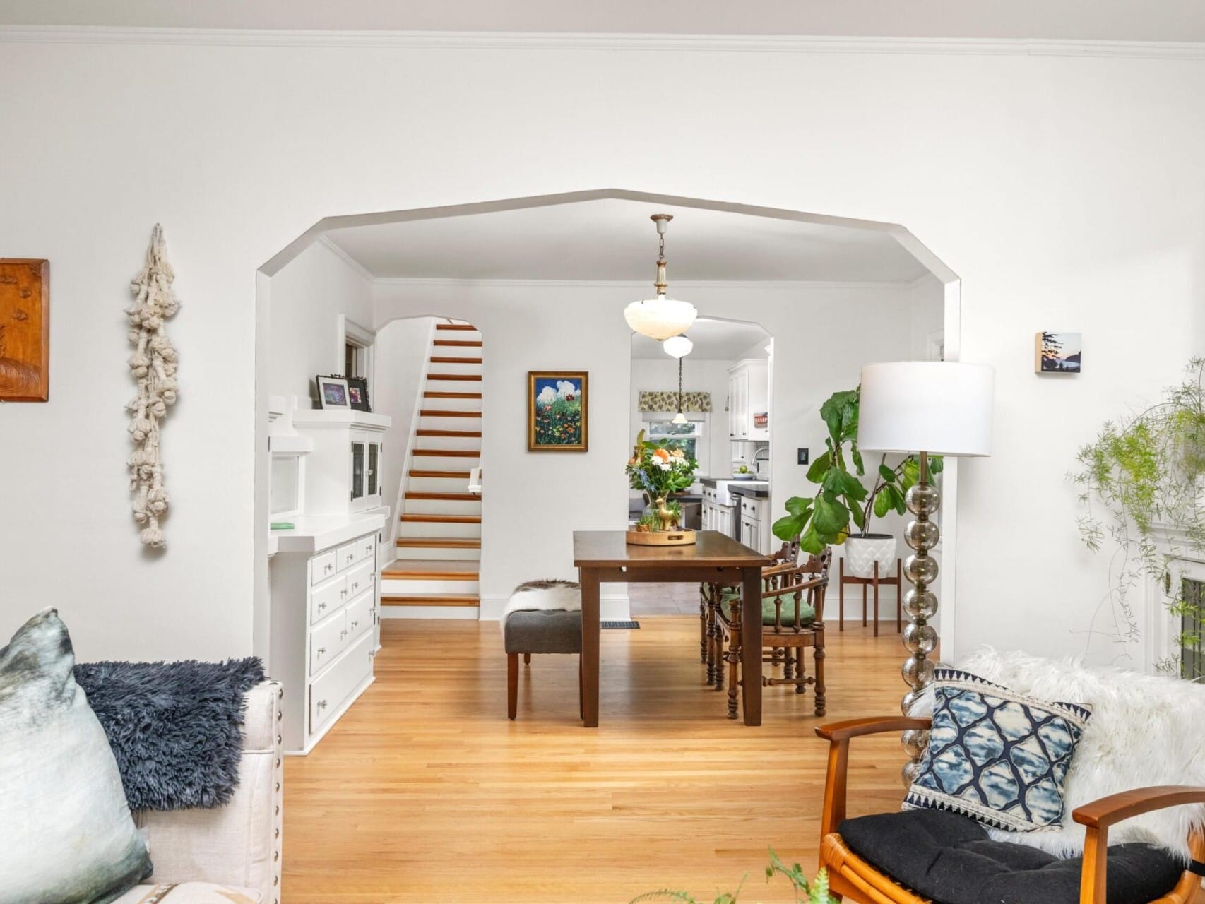 A cozy living room with wooden flooring extends into a dining area, perfect for any Portland Oregon real estate enthusiast. A wooden table with chairs sits under a hanging light. Stairs in the background lead up, while plants, paintings, and white furnishings add charm. A large lamp stands near a fireplace.