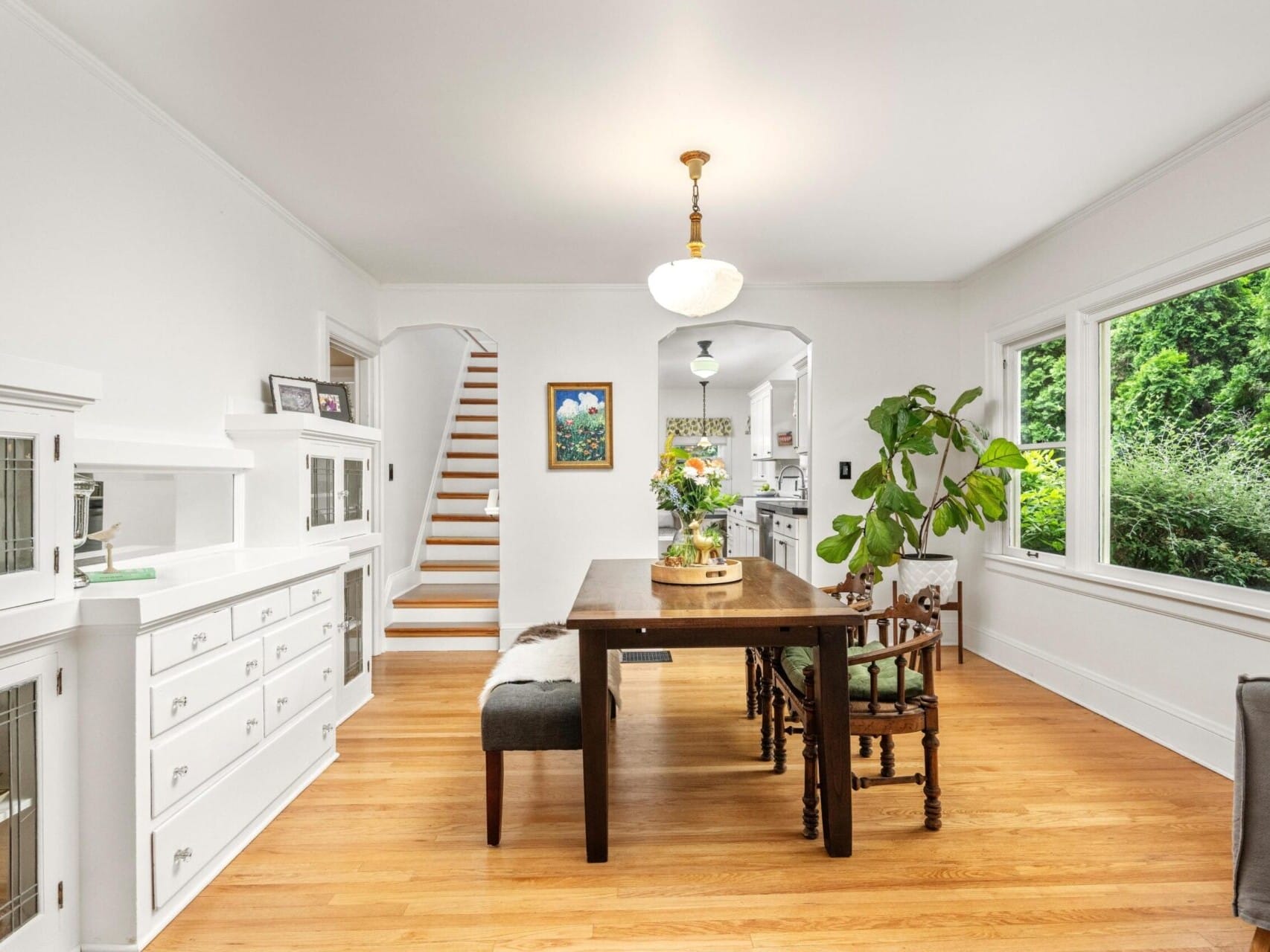 A bright dining room in a charming Portland home features a wooden table and chairs, expansive window views of lush greenery, and crisp white walls. To the left, you'll find a staircase, a plant by the window, and a cabinet with glass doors. Nearby hangs an art piece close to the stairs.