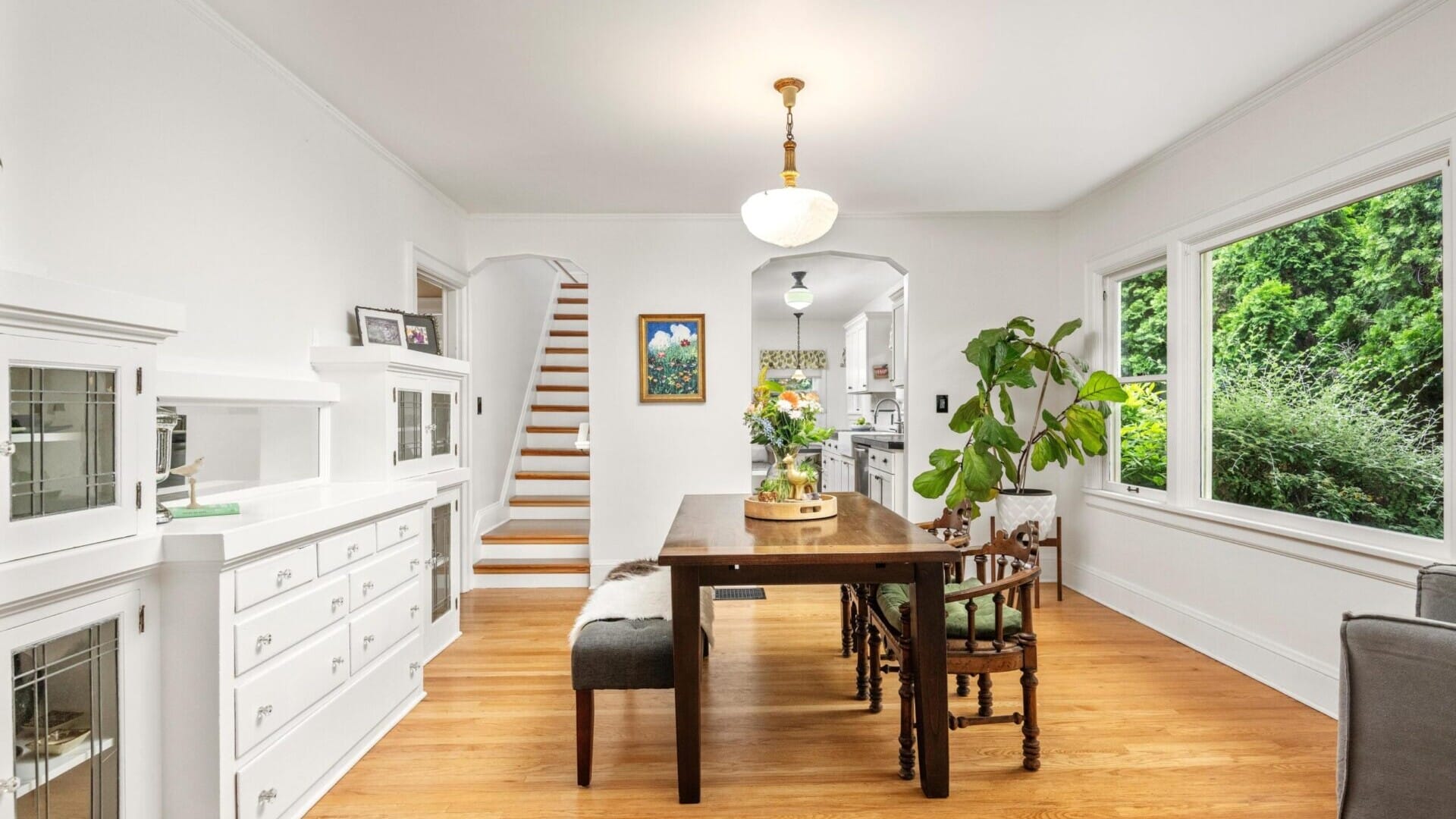 A bright dining room in a charming Portland home features a wooden table and chairs, expansive window views of lush greenery, and crisp white walls. To the left, you'll find a staircase, a plant by the window, and a cabinet with glass doors. Nearby hangs an art piece close to the stairs.