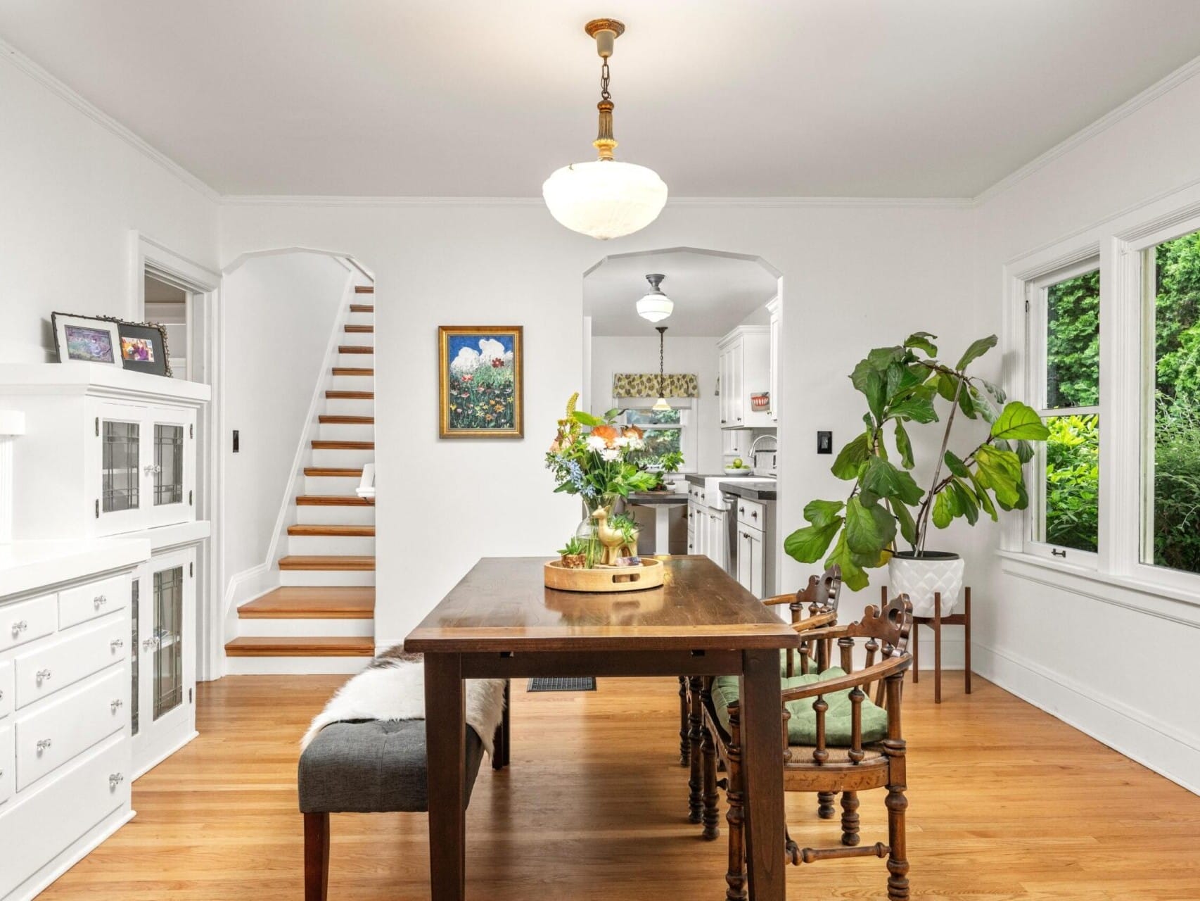 A bright dining area, ideal for Portland Oregon real estate enthusiasts, features a wooden table with a round tray of plants, surrounded by chairs. A cushioned bench sits to one side. A staircase is visible on the left, and large windows offer a view of greenery outside. The space is airy and inviting.