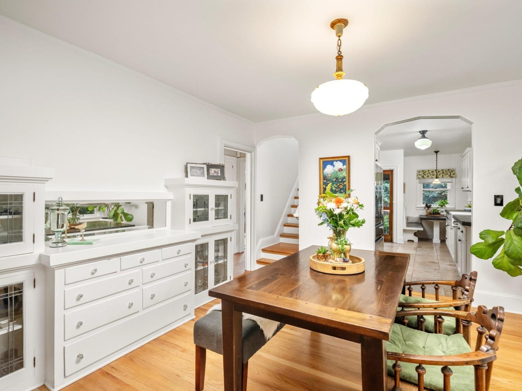 A bright dining room with a wooden table and two chairs invites you in. White cabinets with glass doors adorn the wall, reflecting the charm of Portland Oregon real estate. A potted plant adds life, while a pendant light casts a warm glow. Stairs lead to a kitchen view in the background.