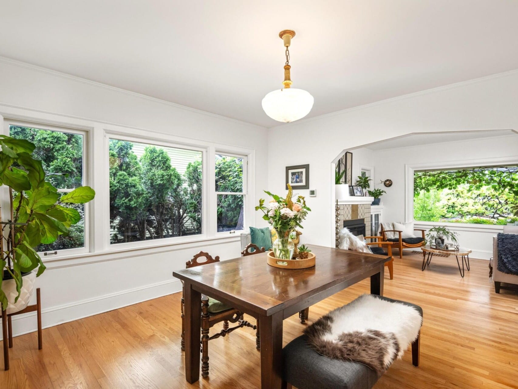 A bright dining room with a wooden table and chairs, adorned with a vase of flowers, is what you will find in this charming piece of Portland Oregon real estate. Large windows overlook lush greenery, complementing the cozy living area with a fireplace and art. A chandelier beautifully graces the ceiling.