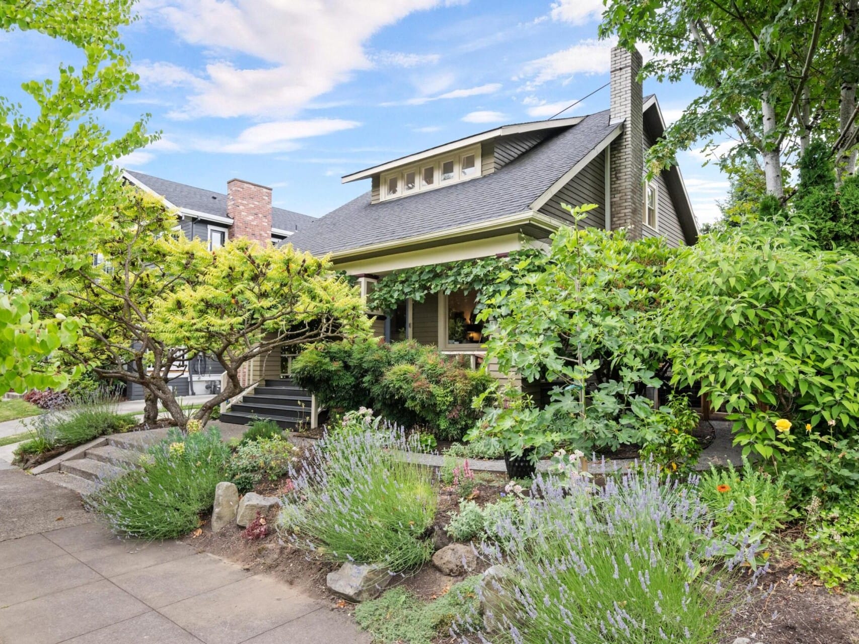 A charming house with a gabled roof, surrounded by lush greenery and flowering plants, embodies the Portland Oregon real estate dream. A pathway leads to the entrance, flanked by bushes and trees. The clear sky adorned with a few clouds completes this peaceful suburban scene.