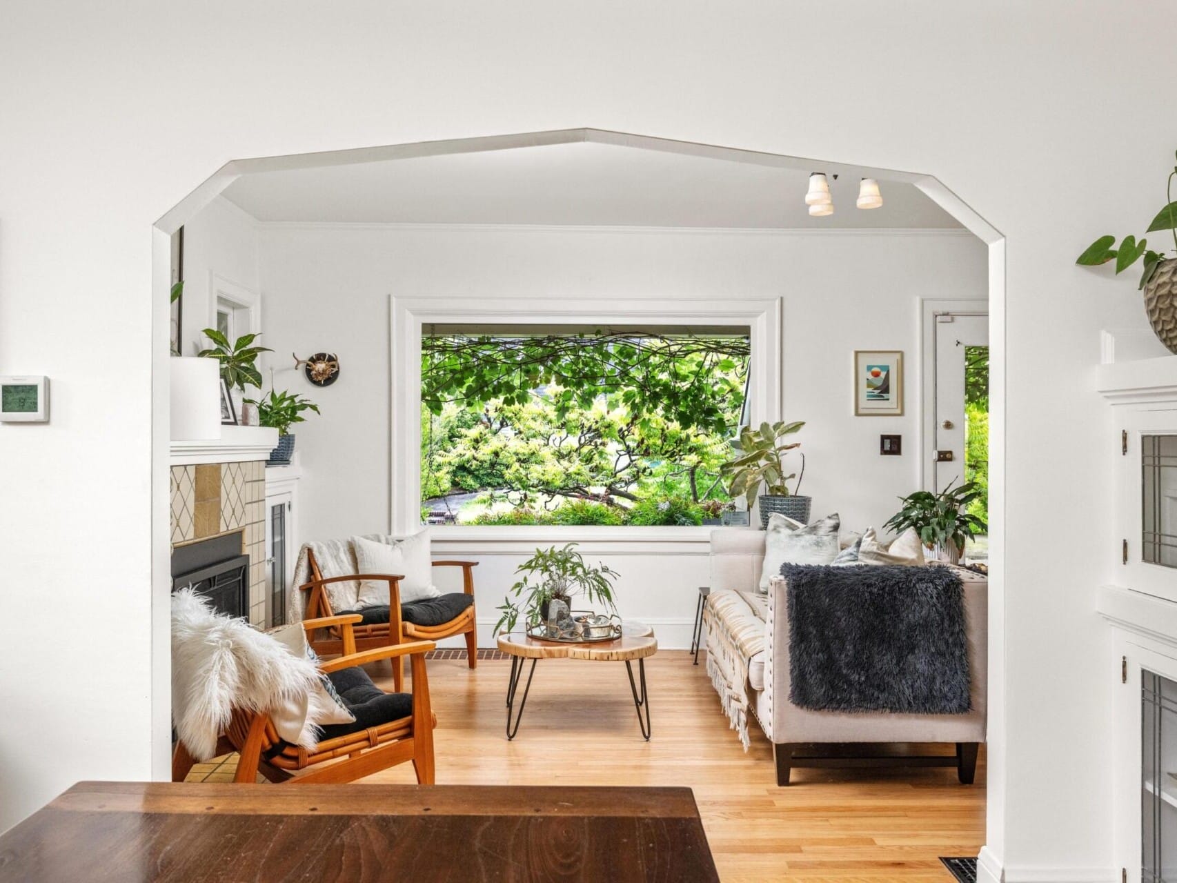 A cozy living room with wooden flooring features a fireplace, two armchairs with cushions, a daybed, and a large window overlooking lush greenery. Plants and framed pictures add to the decor, creating a serene atmosphere—a perfect find for anyone exploring Portland Oregon real estate.