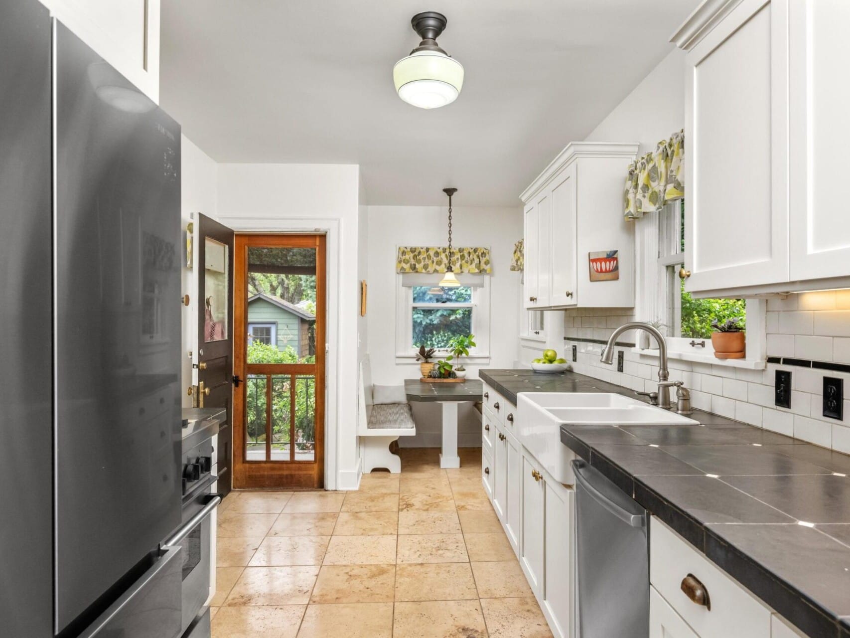 A modern kitchen in a sought-after Portland, Oregon real estate listing boasts white cabinets, black countertops, and beige tile flooring. A farmhouse sink faces the window beside a stainless steel fridge. Sunlight streams through the glass door leading to a garden and cozy breakfast nook.