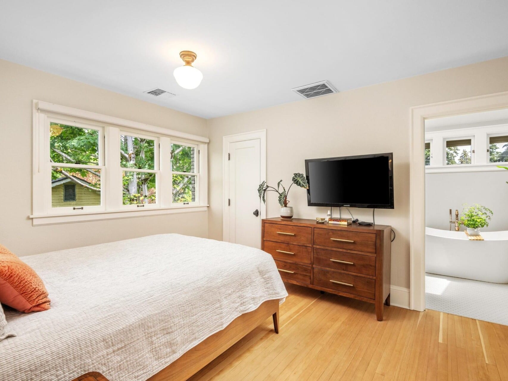 A bright bedroom in Portland Oregon Real Estate showcases a bed with patterned pillows, a wooden dresser with a TV on top, and a door leading to a bathroom with a freestanding bathtub. Large windows flood the wooden floor with natural light.