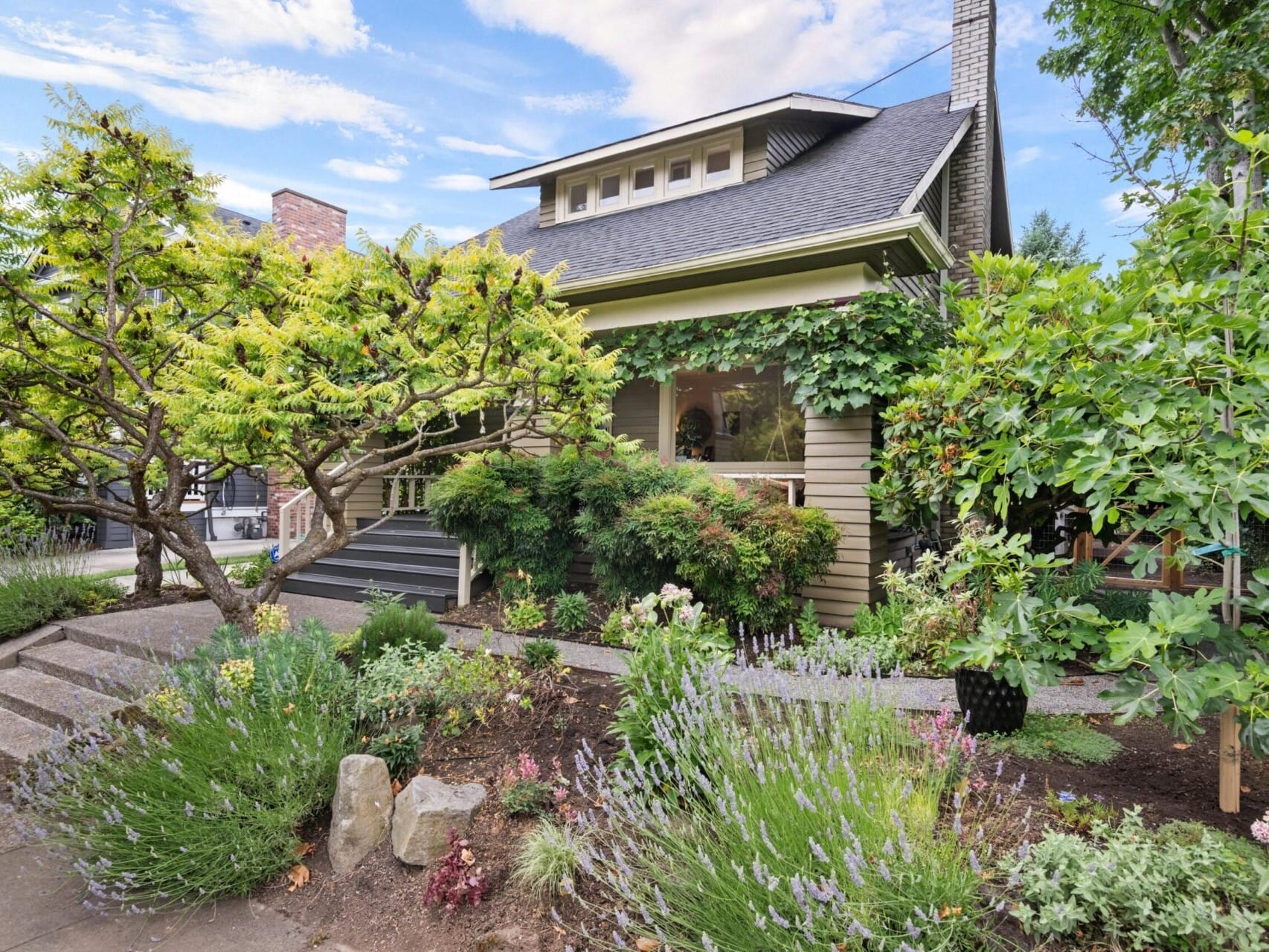 A charming house in Portland, surrounded by lush greenery and flowering plants, captures the essence of Portland Oregon real estate. Featuring a mix of shrubs and trees with a wooden porch partially covered by vines, a pathway leads to the front steps, creating a welcoming garden setting.