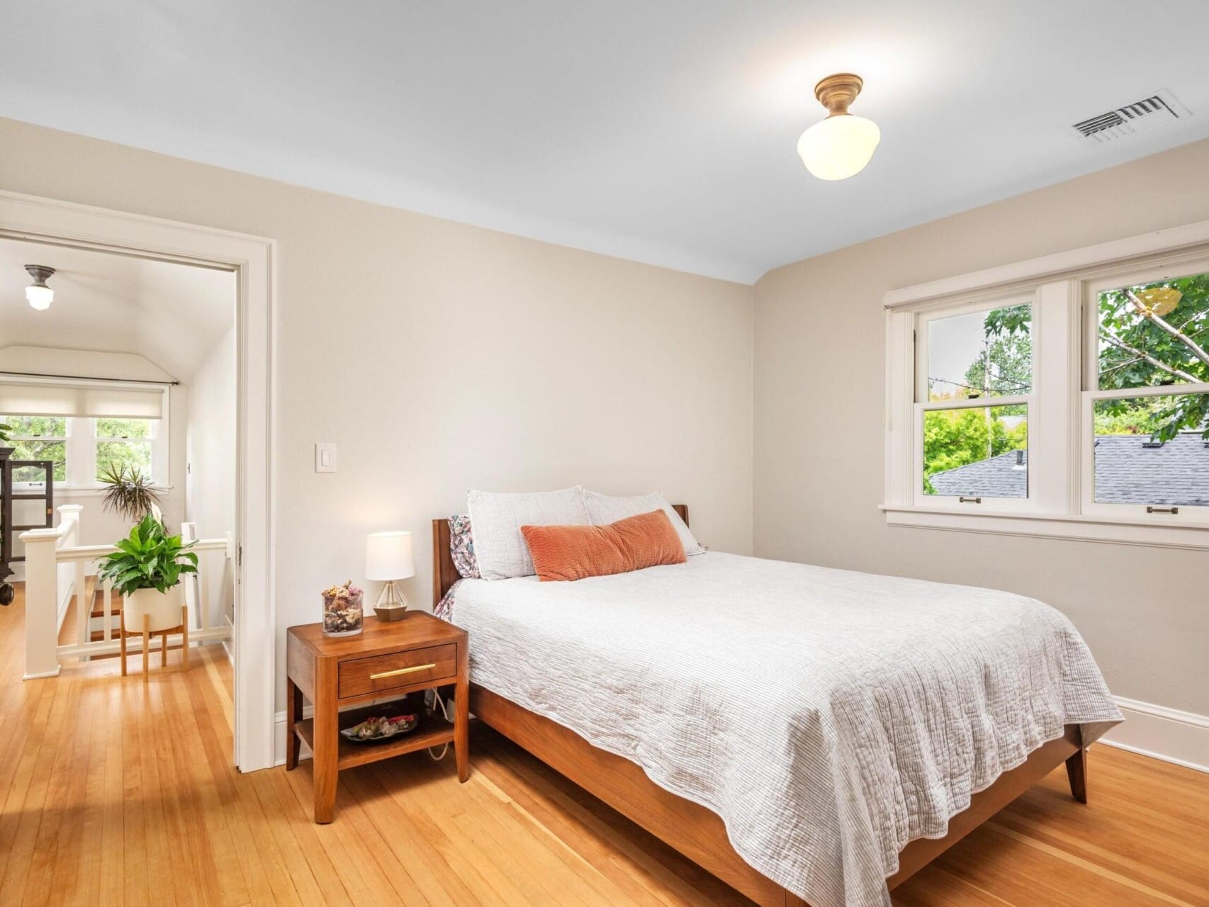 A cozy bedroom with a wooden floor, featuring a neatly made bed with white and orange bedding. A wooden nightstand with a lamp and decor sits beside the bed, curated by a top Portland Realtor. A window offers a view of greenery, and an open door leads to another room.