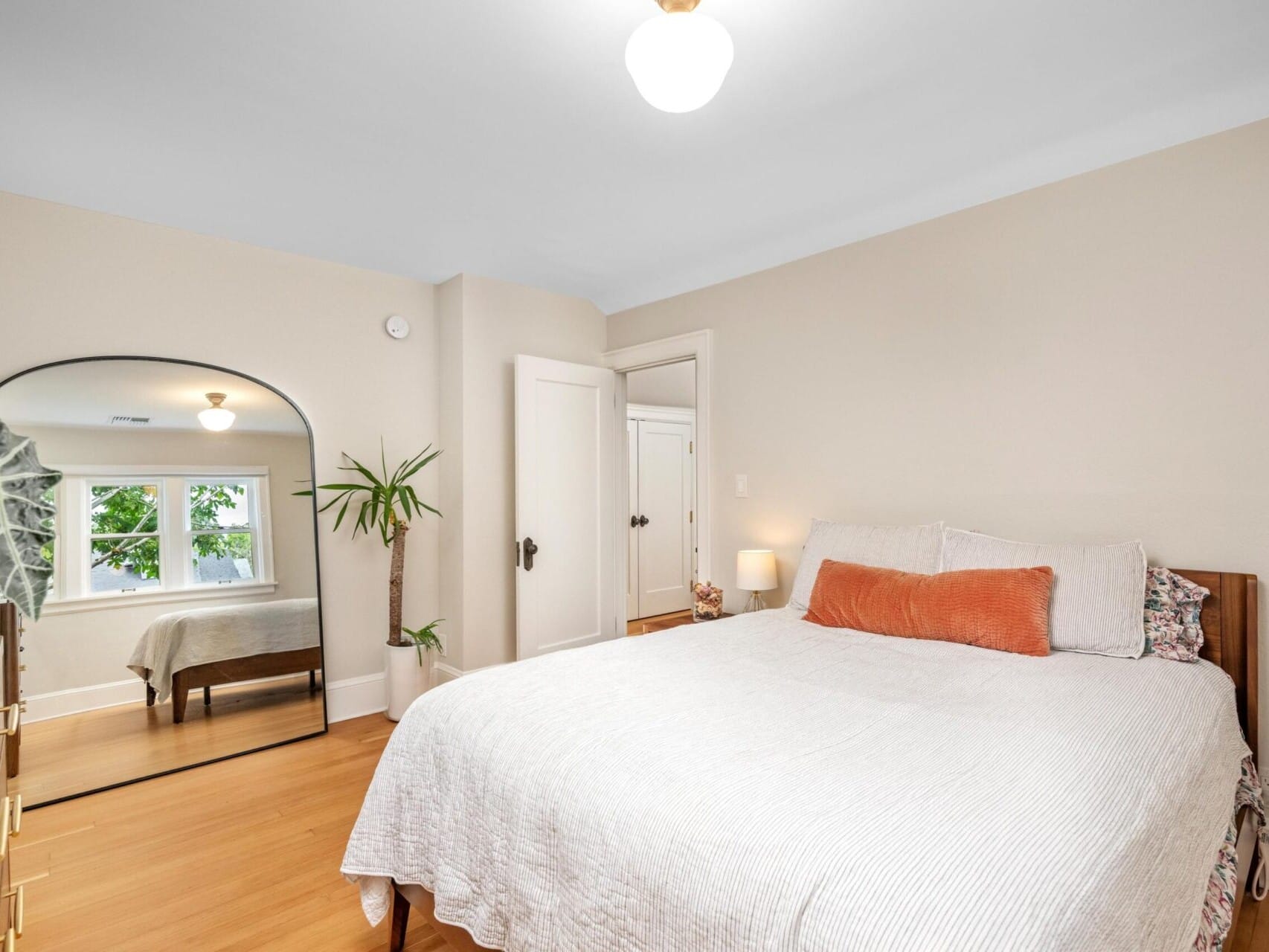 A cozy bedroom in a charming Portland Oregon real estate listing features a neatly made bed with white linens and an orange accent pillow. A tall plant stands by a large, arched mirror next to the window. A wooden dresser is on the left, while soft lighting illuminates the room.