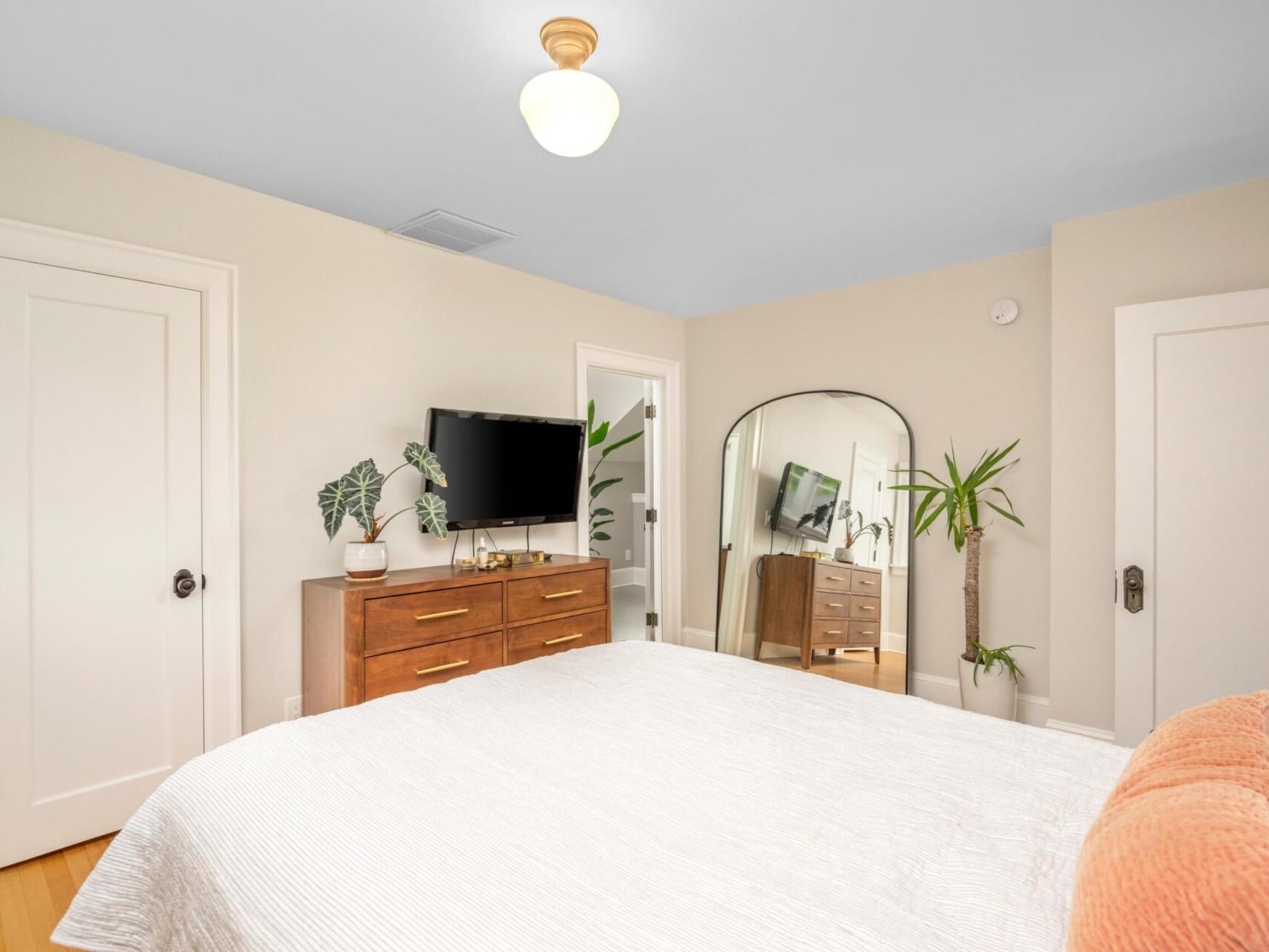 A bright bedroom in a Portland Oregon real estate gem features a large bed with white bedding and a peach cushion. A wooden dresser with a TV adorns the wall, complemented by a tall mirror and potted plants. The room's white walls, wooden floors, and ceiling light showcase its elegant charm.