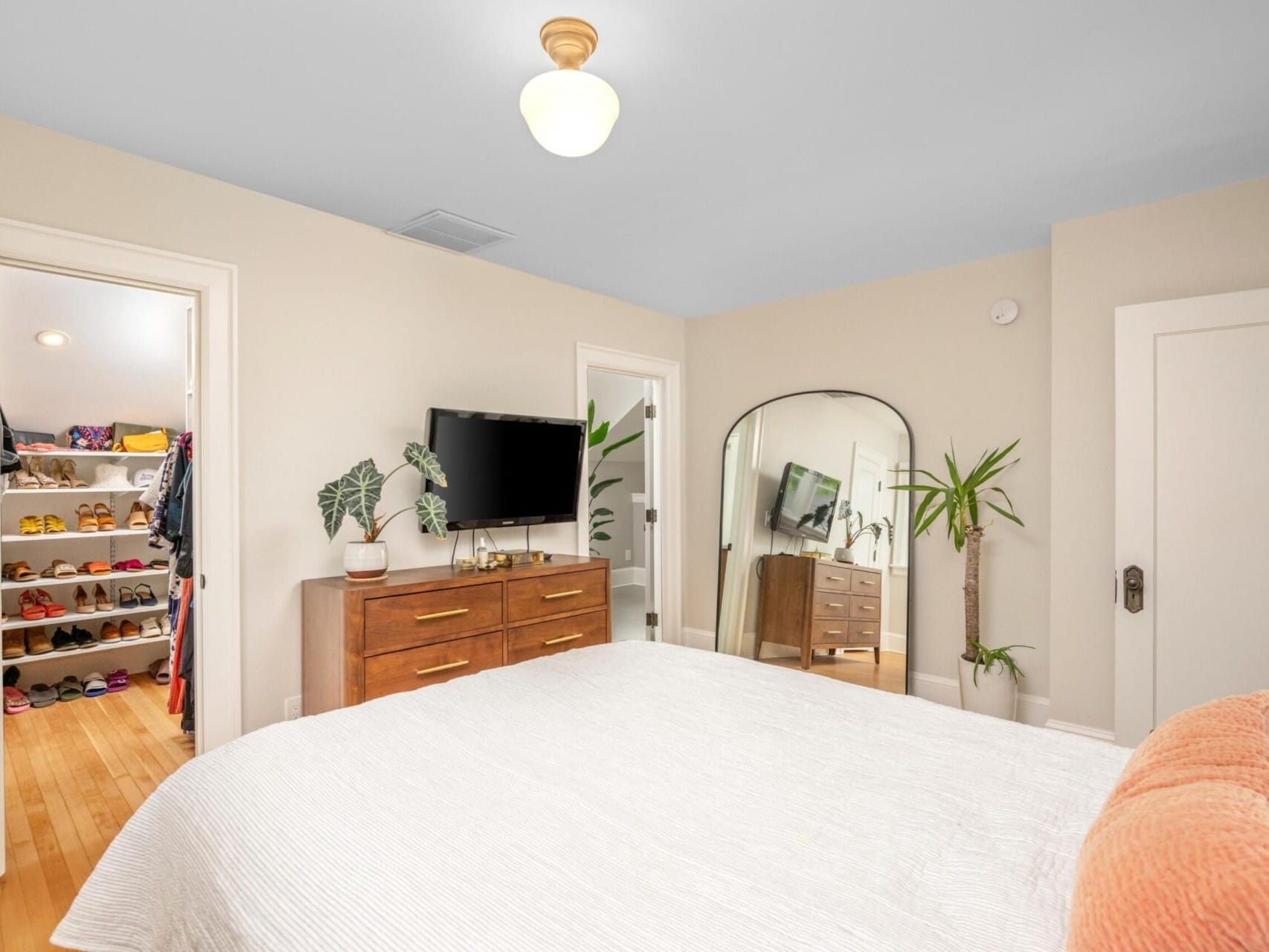 Discover this dreamy Portland Oregon real estate listing featuring a cozy bedroom with beige walls and a large bed adorned in white and peach bedding. The space includes a curved mirror, wooden dresser with TV, plants, and an open door leading to a walk-in closet filled with shoes.