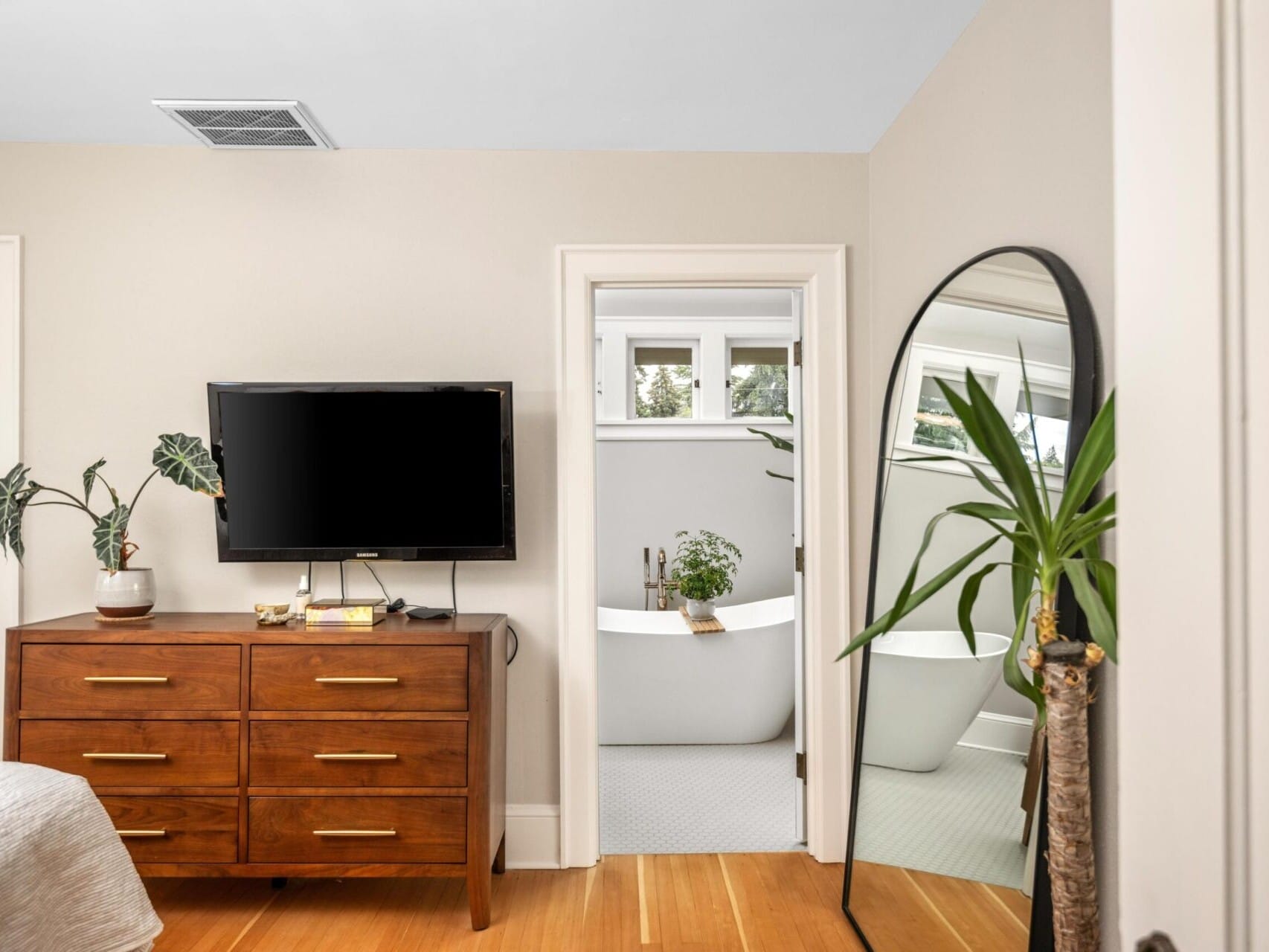 A bedroom owned by a Portland Realtor, featuring a wooden dresser, TV, plants, and a large arched mirror. An open door reveals a bathroom with a modern white bathtub and greenery. Warm, neutral tones create a cozy atmosphere.