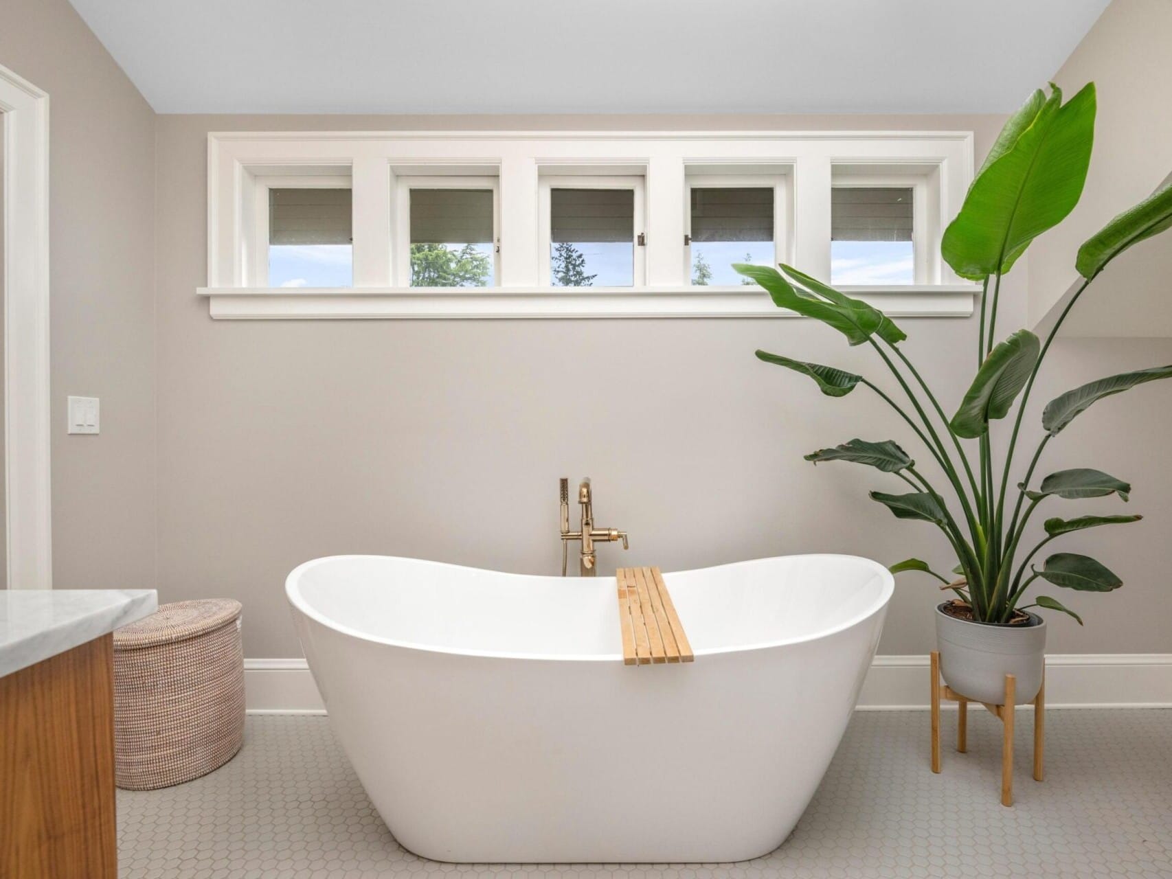 A modern bathroom in a Portland Oregon real estate gem, featuring a freestanding white bathtub with a wooden bath tray. A large green plant rests beside the tub, while small windows above let in natural light. There's a woven basket near the door, and neutral-toned walls complement the light gray floor.