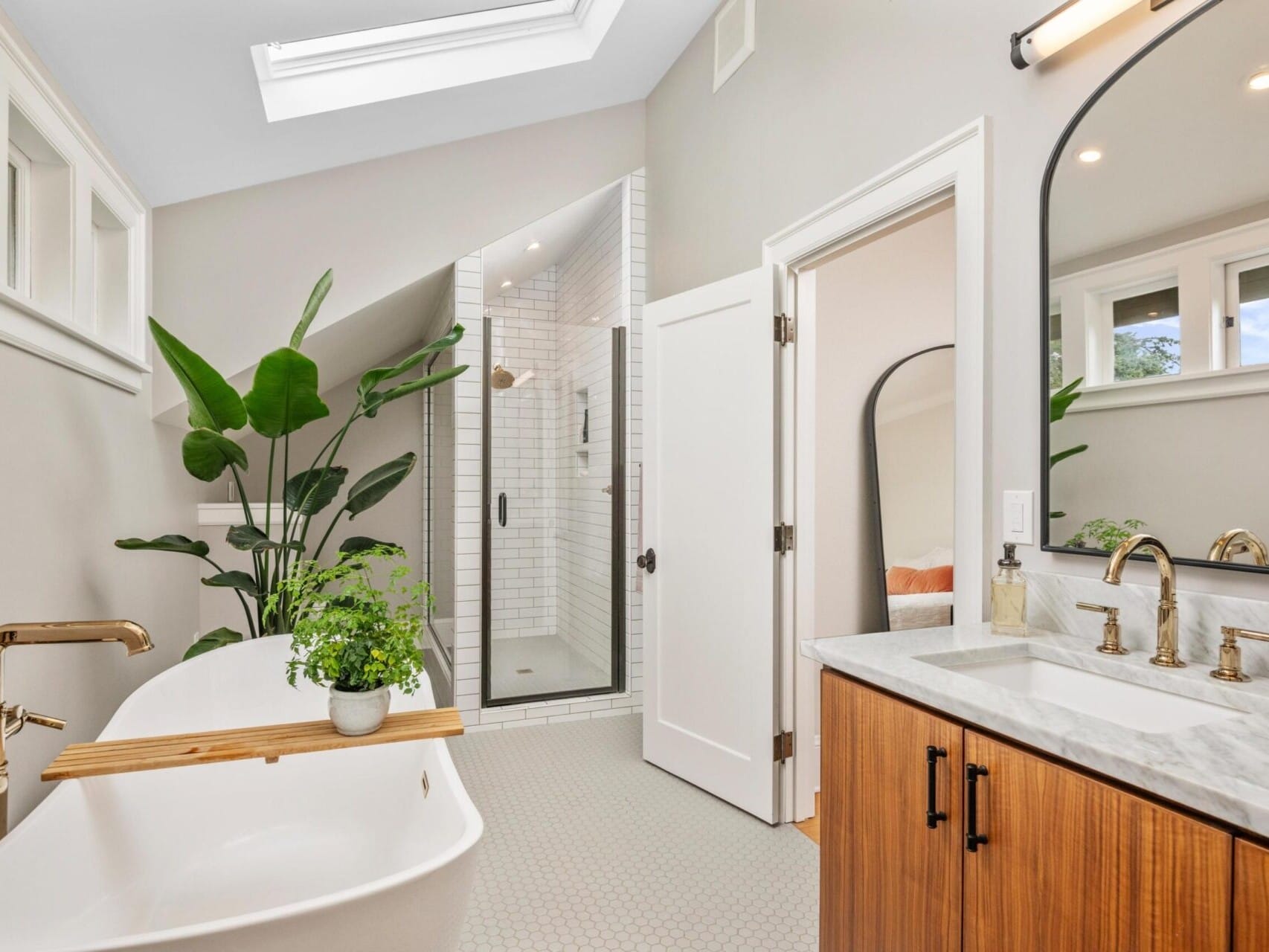 This modern bathroom, found in an exquisite Portland Oregon real estate gem, features a freestanding white bathtub with a wooden tray adorned with plants, a glass-enclosed shower, and a large mirror above a marble-topped vanity. A skylight and small windows illuminate the space with fresh greenery.