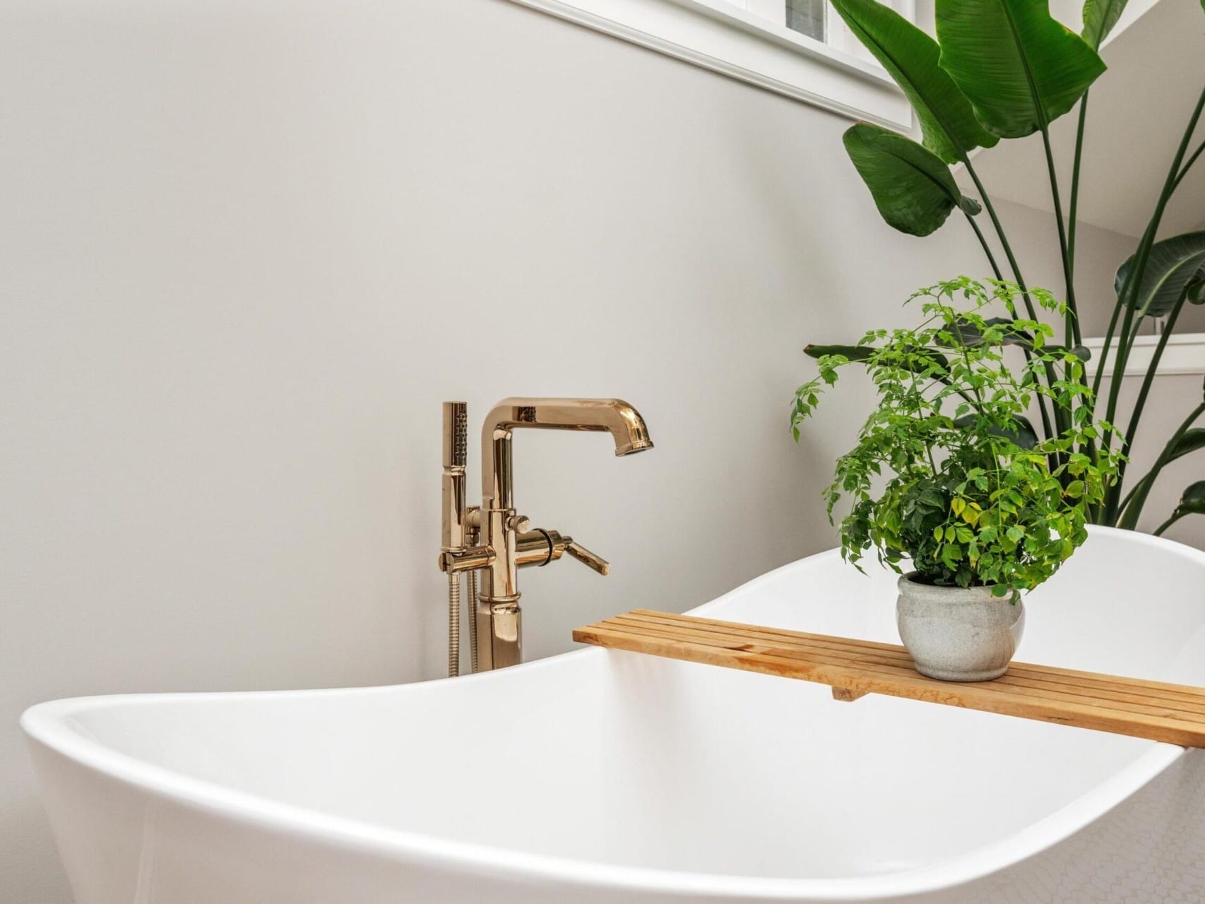 A modern bathroom, ideal for Portland Oregon real estate enthusiasts, features a white freestanding tub with a gold faucet. A wooden tray rests across the tub, holding a small potted plant. A large green plant stands beside the tub against a light gray wall, creating a serene atmosphere.