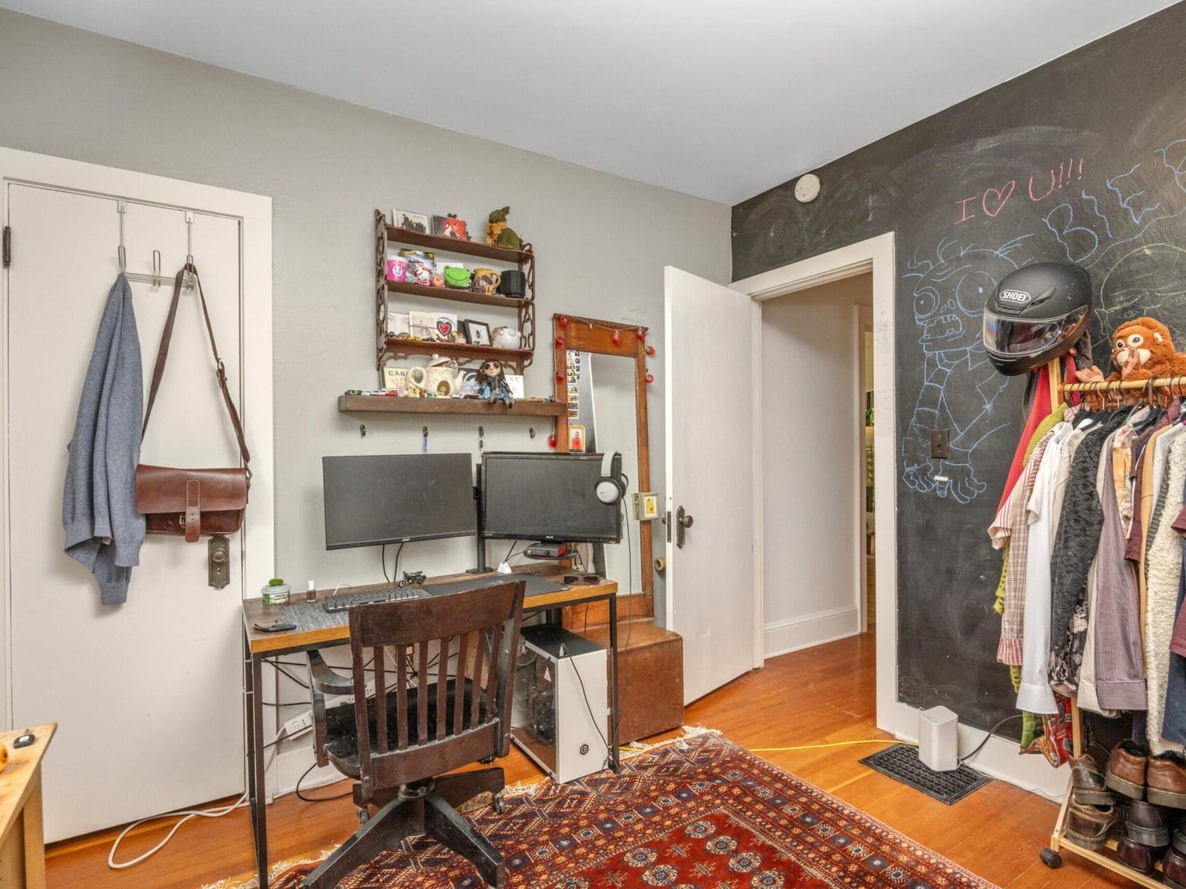 A cozy room perfect for a Portland realtor, featuring a desk with dual monitors and a wooden chair. Shelves with various items are above the desk. A chalkboard wall on the right displays drawings and writing. The floor is hardwood with a patterned rug, and a bag hangs on the door.