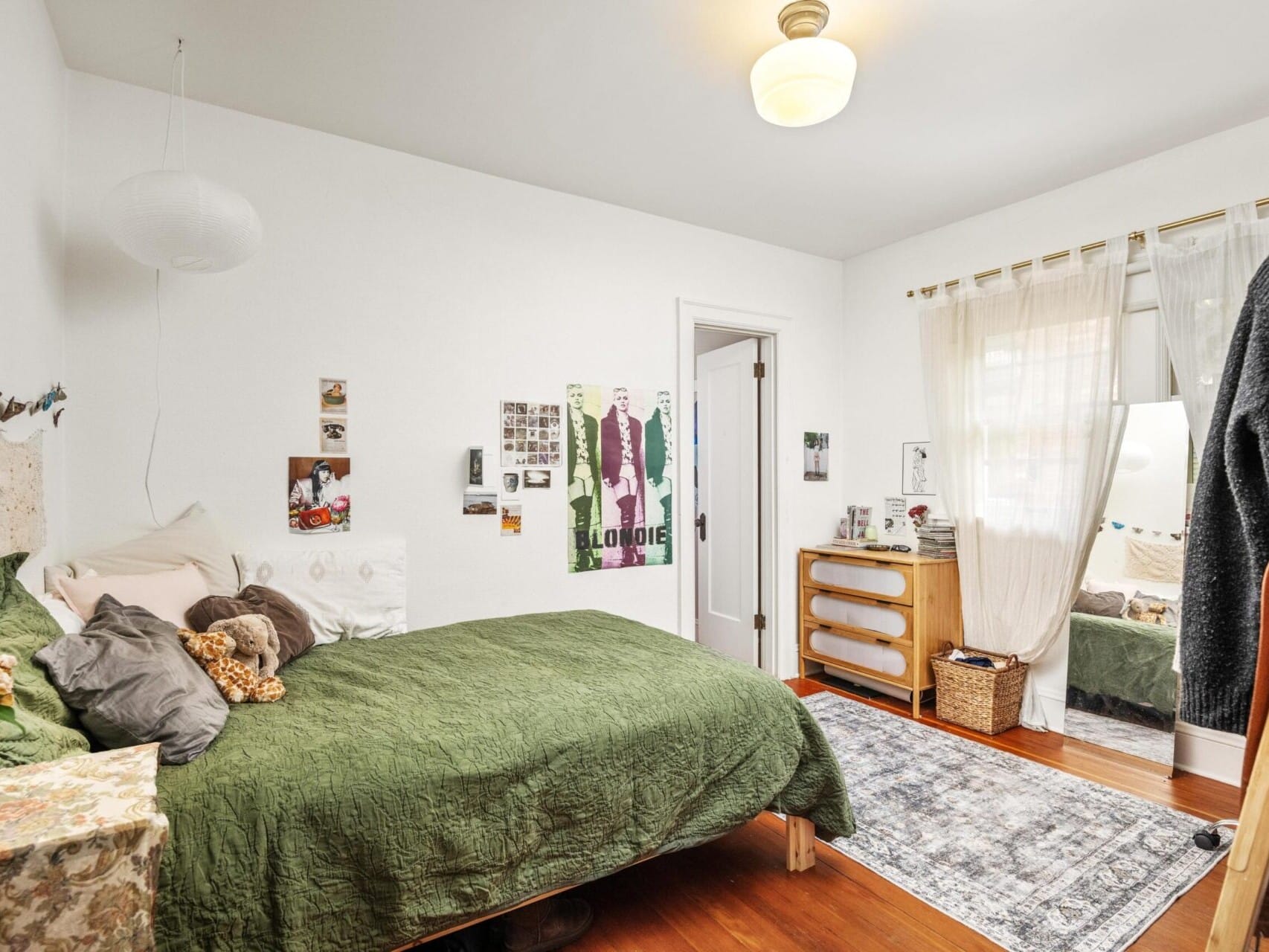 A cozy bedroom in a charming Portland home features a green bedspread, pillows, and a small teddy bear on the bed. Posters adorn the white walls alongside a wooden dresser with a mirror. The room boasts hardwood floors, a light rug, and sheer curtains—a gem in Portland Oregon Real Estate.