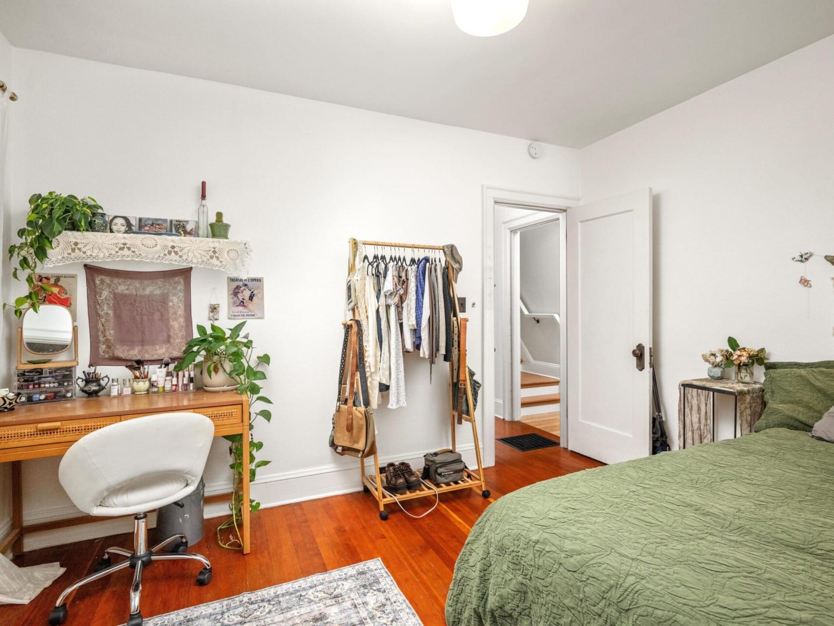 A cozy bedroom in Portland, featuring white walls and a wooden floor. A bed with a green quilt sits next to a small wooden vanity adorned with plants, while a clothes rack holds hanging garments. A white chair and decorative rug complete this inviting space, listed by the top real estate agent in Portland.