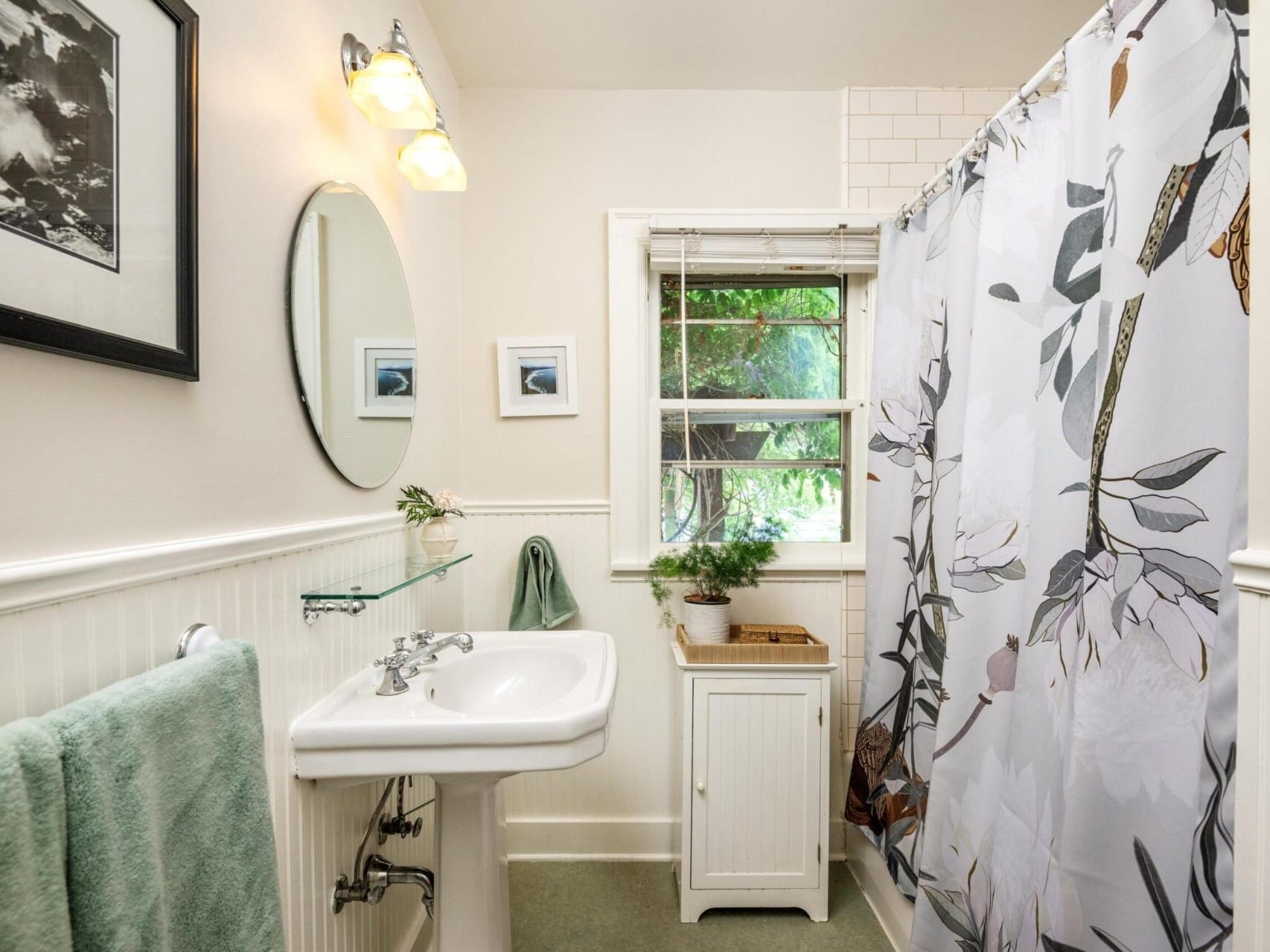 A small bathroom showcased by a top real estate agent features a white sink beneath a round mirror, a towel on a rack, and a potted plant on the cabinet. The shower's floral curtain adds charm. A partially open window reveals Portland Oregon's greenery, with framed photos adorning the walls.