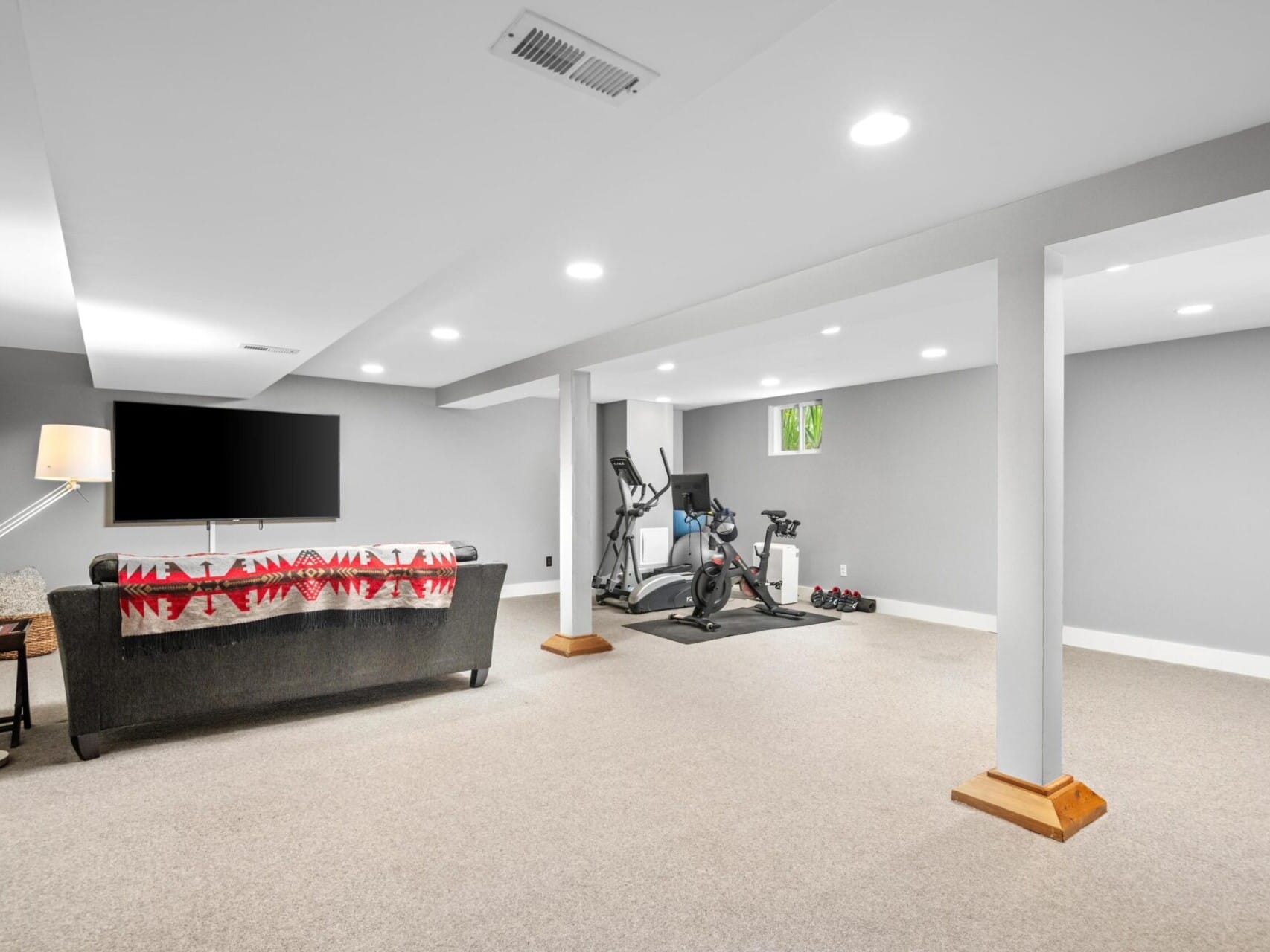 A spacious basement in Portland, Oregon, with gray walls features a couch with a red-patterned throw, a large flat-screen TV, and exercise equipment including two stationary bikes and weights. Warm lighting and carpeted flooring create a cozy atmosphere ideal for any homeowner.