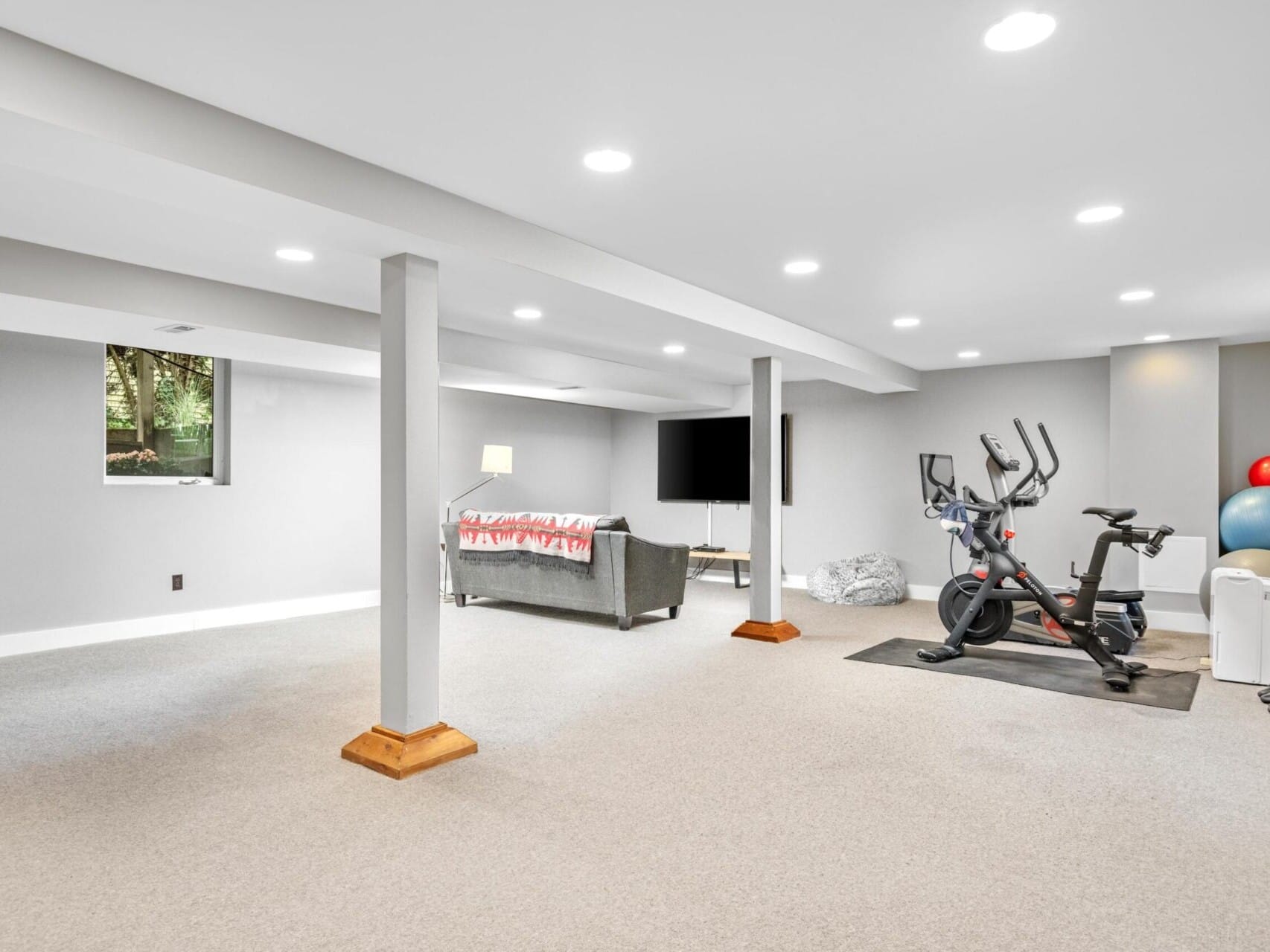 A spacious basement room with a gray carpeted floor features an exercise area equipped with a stationary bike and weights. Nearby, a cozy sitting area includes a gray sofa facing a large flat-screen TV. Recessed ceiling lights illuminate this ideal Portland Oregon real estate find.