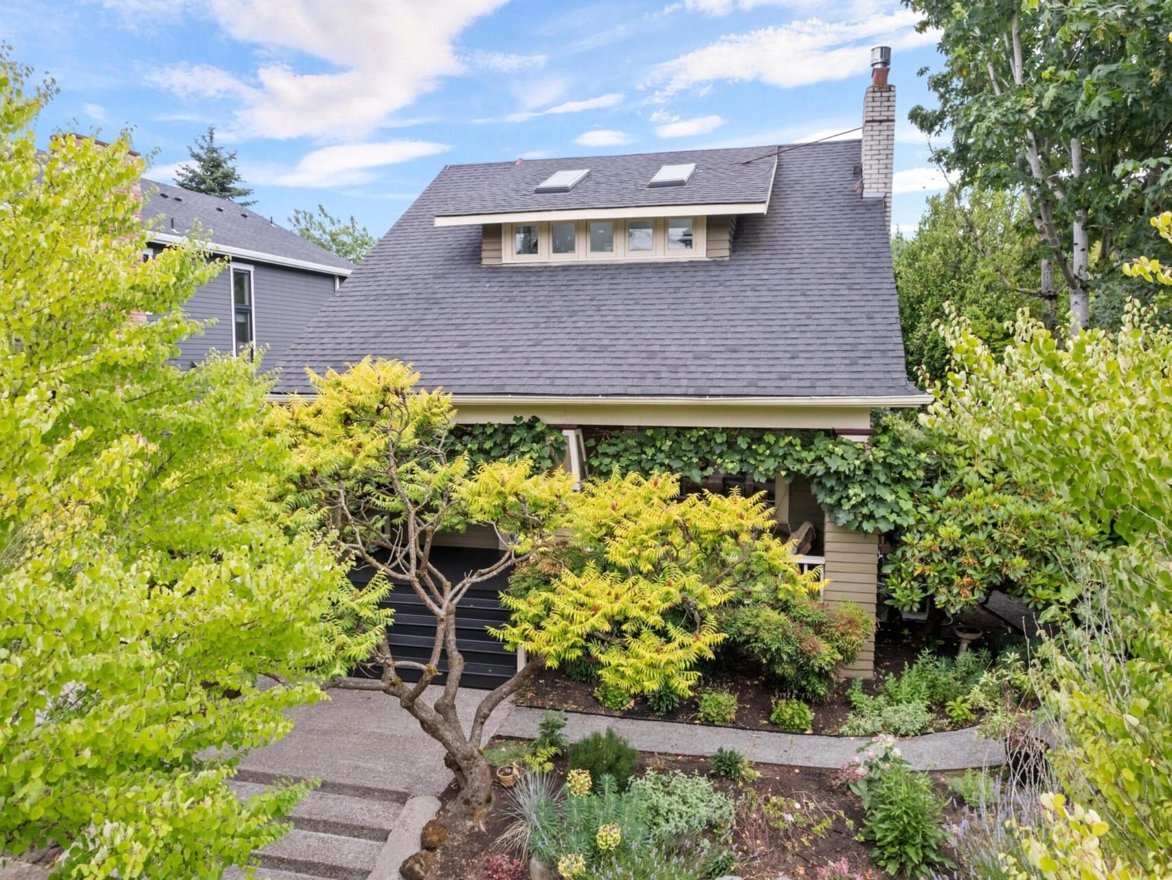 Two-story house with a dark roof and covered porch, nestled amidst lush green trees and plants in beautiful Portland, Oregon. A winding path leads to the entrance under a partly cloudy sky—ideal for those exploring Portland Oregon real estate's exquisite charm.